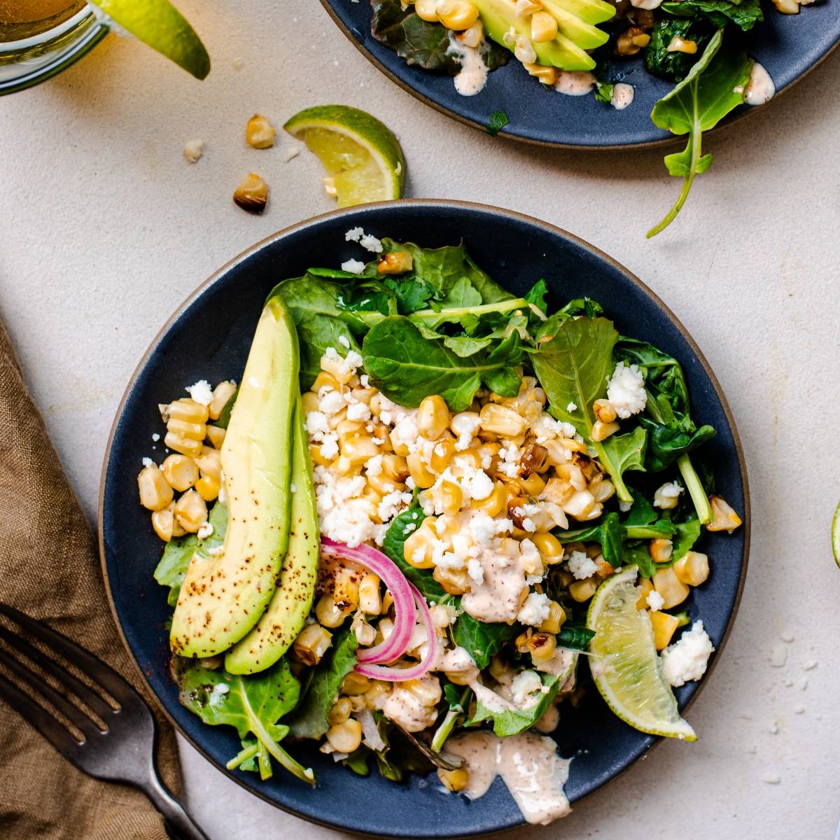 A vibrant Elote Corn Kale Salad with leafy greens, grilled corn, avocado slices, pickled onions, cheese, and lime on a black plate—all brought together with a spicy yogurt lime dressing.