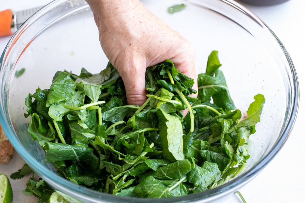 A hand mixing fresh leafy greens in a clear glass bowl, preparing a vibrant Elote Corn Kale Salad with spicy yogurt lime dressing.