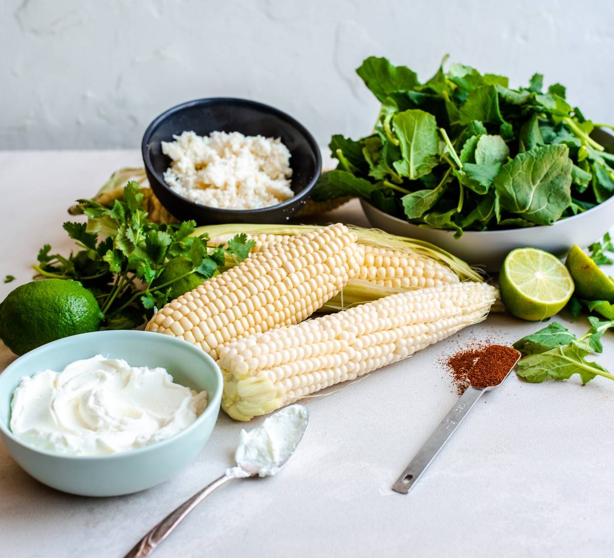 Fresh ingredients including corn, leafy greens, cheese, lime, chili powder, cilantro, and a bowl of cream come together in this Elote Corn Kale Salad with a Spicy Yogurt Lime Dressing.