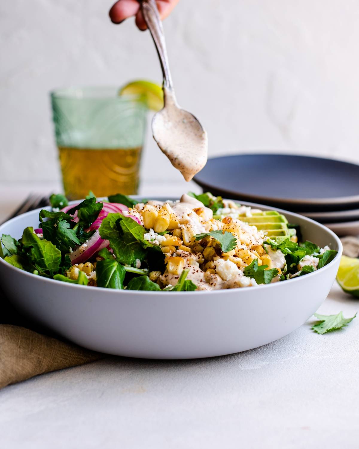 A hand drizzles spicy yogurt lime dressing over a fresh salad in a white bowl, with a drink and plates in the background—perfect for elevating your kale salad recipe.