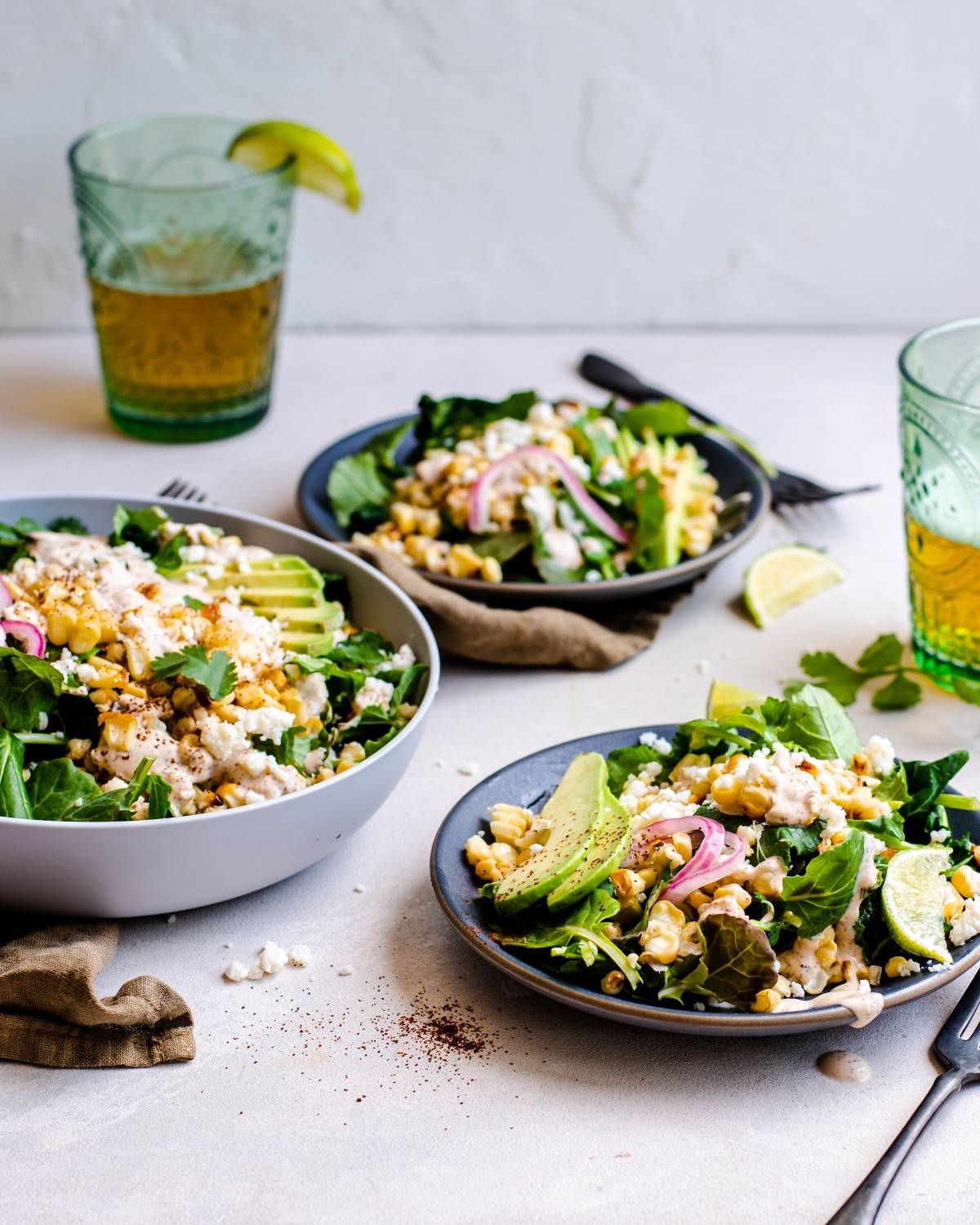 Mexican inspired salad with greens, avocado, corn, and cheese on plates and in a bowl, served with drinks, lime wedges, and a spicy yogurt lime dressing.
