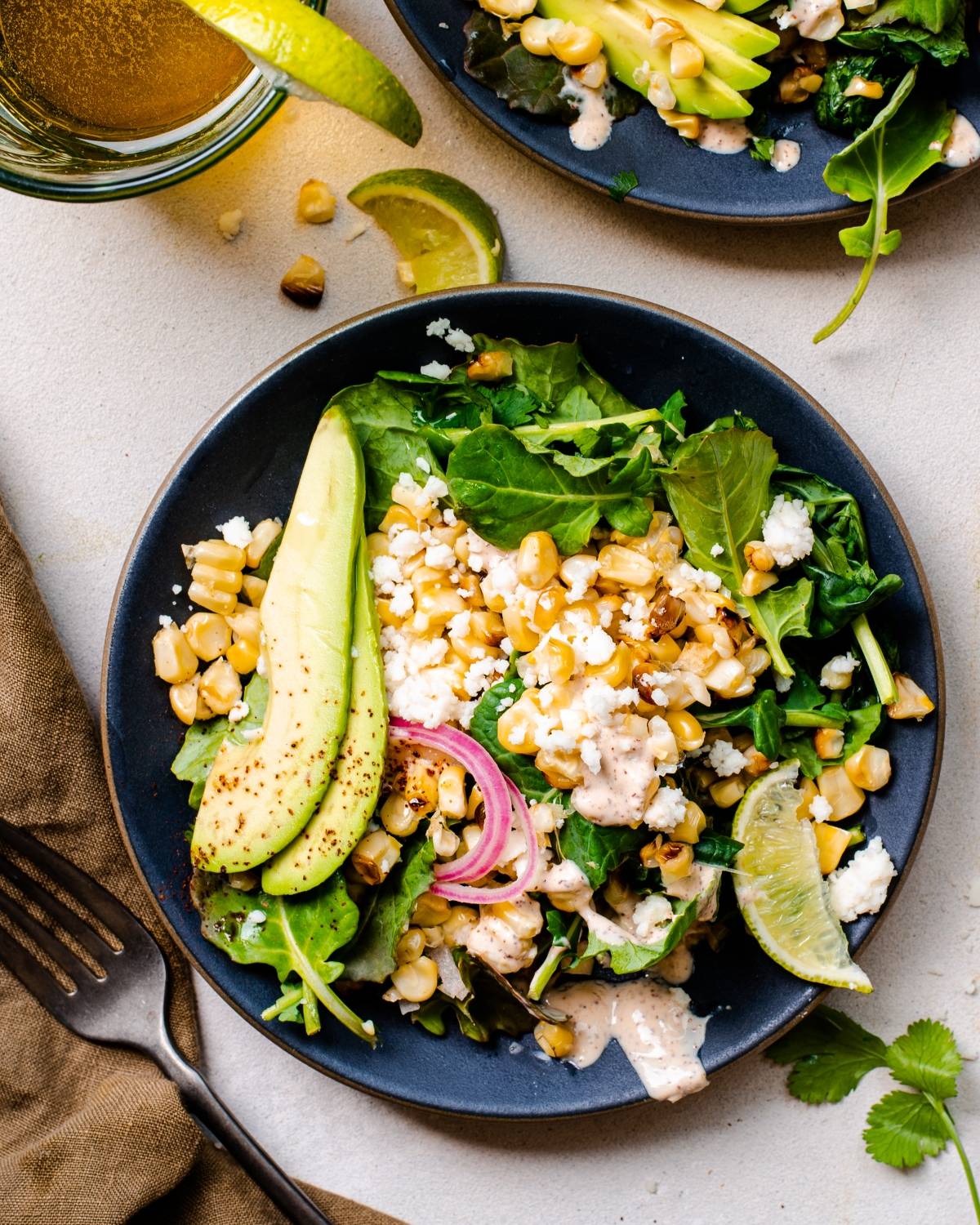 A Mexican inspired salad with avocado, corn, greens, pickled onions, cheese, and lime on a dark plate, finished with a spicy yogurt lime dressing.