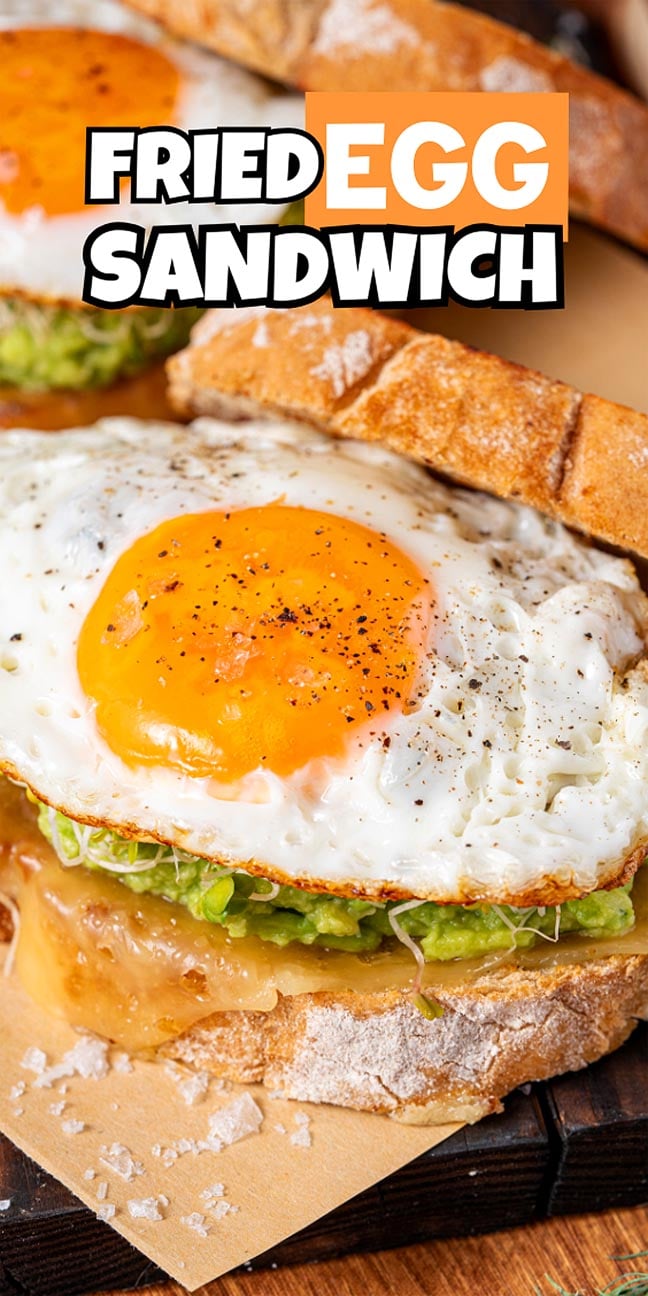 Close-up of a fried egg sandwich with creamy avocado on rustic bread, topped with pepper and melted cheese for the ultimate breakfast bite.