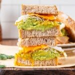Stacked fried egg sandwich halves with avocado, cheese, sprouts, and egg on whole grain bread, served on parchment paper.