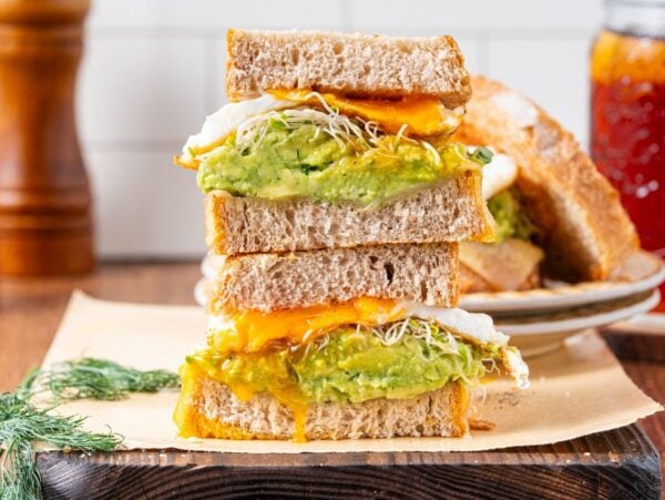 Stacked fried egg sandwich halves with avocado, cheese, sprouts, and egg on whole grain bread, served on parchment paper.