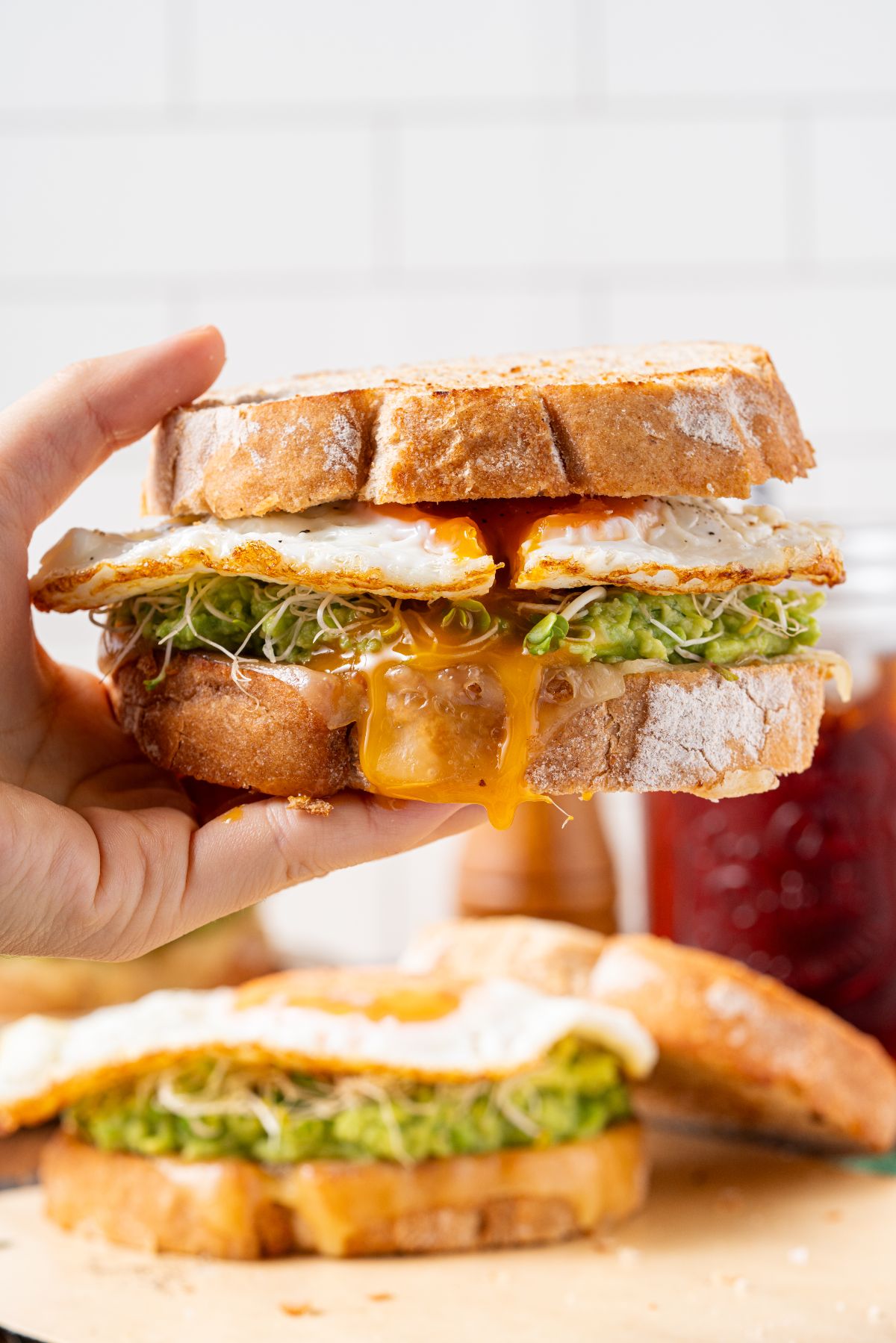 A hand holds a delicious fried egg sandwich with melted cheese, sprouts, and avocado on rustic bread.