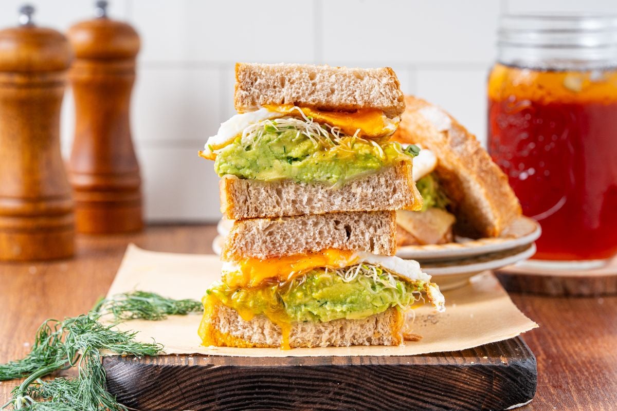 Stacked fried egg sandwich with avocado, sprouts, and melted cheese on whole grain bread, served next to iced tea.