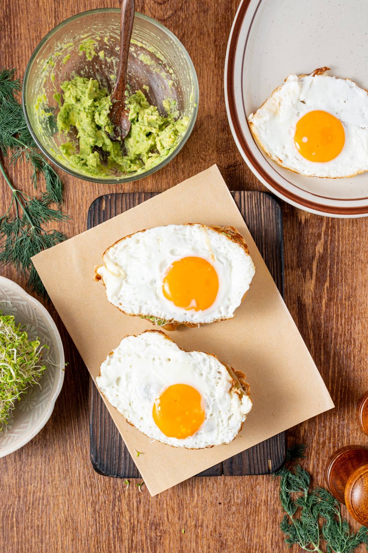 Two slices of bread topped with sunny-side-up eggs, next to a bowl of avocado spread and a plate with another egg.