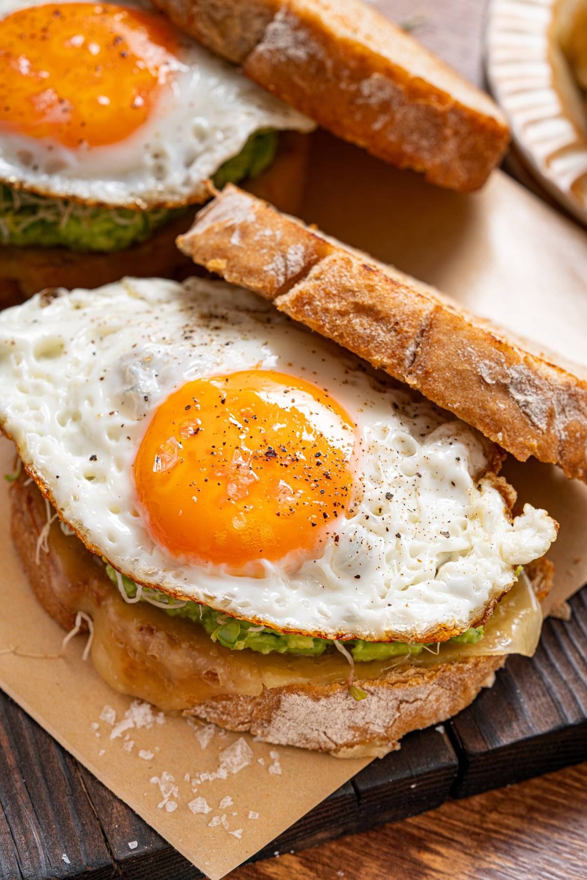 A sandwich with fried egg, avocado, and melted cheese on rustic bread, sprinkled with pepper and salt.