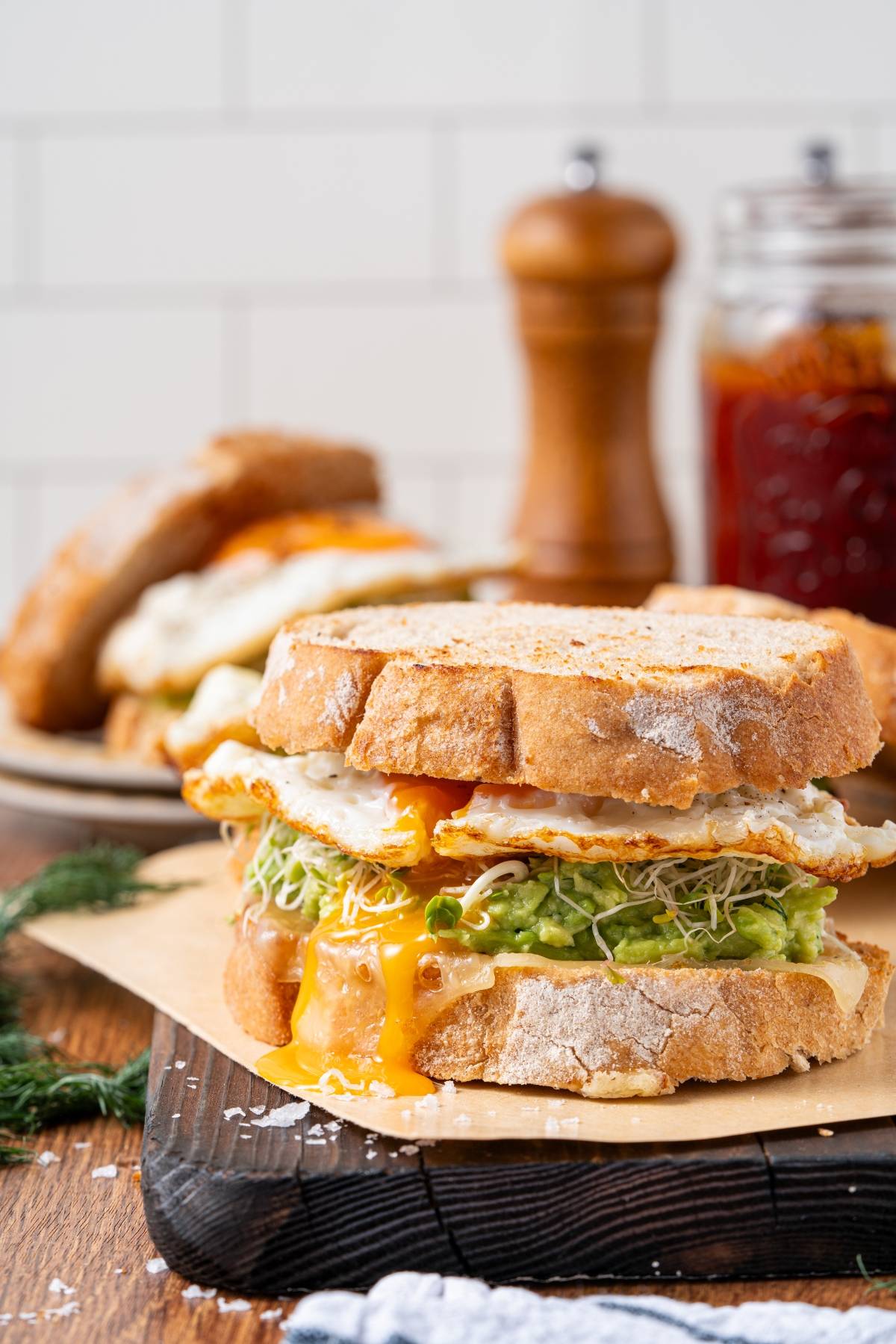 A sandwich with fried egg, avocado, sprouts, and melted cheese on rustic bread, with yolk dripping out.