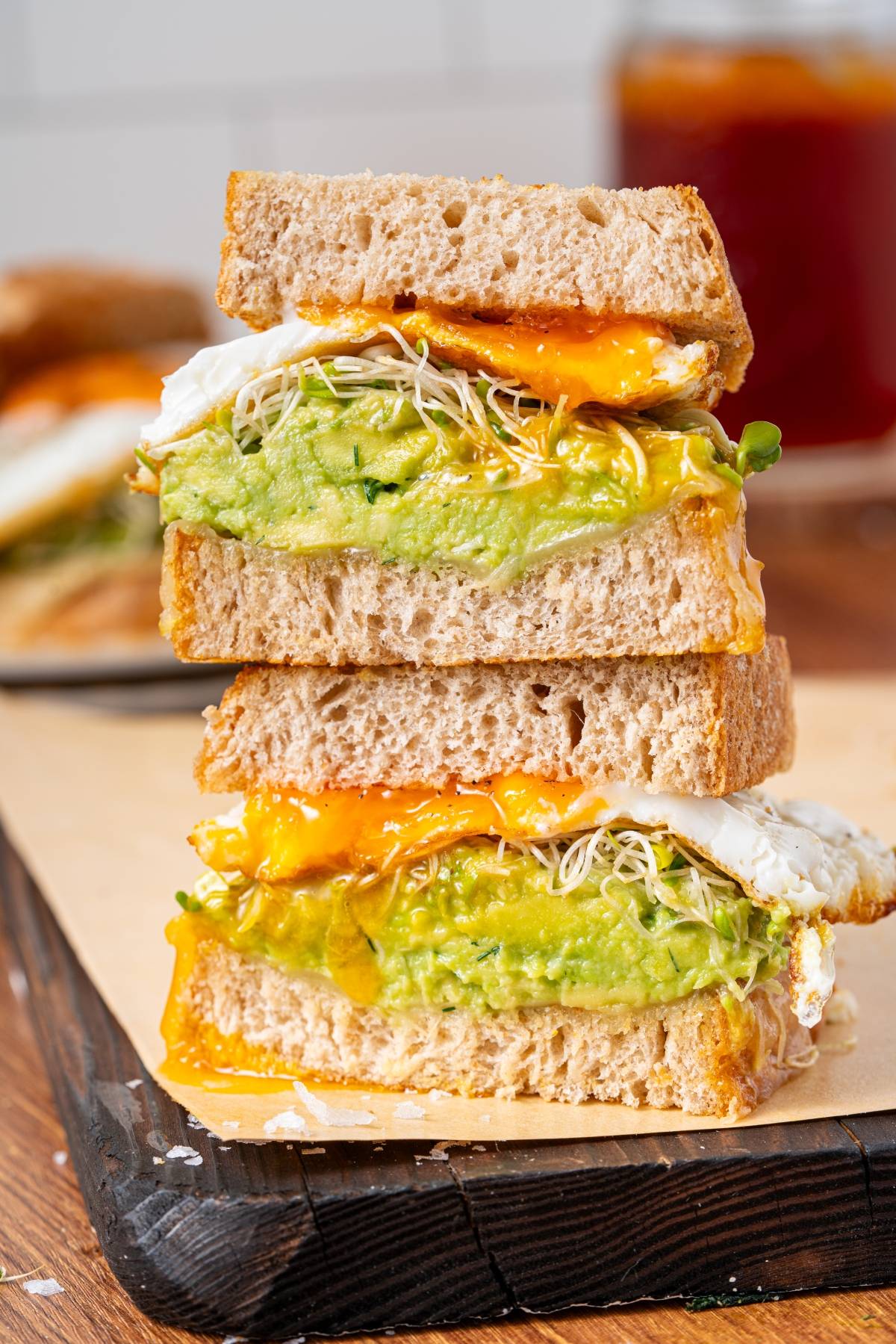Two stacked sandwiches with fried eggs, avocado, and sprouts on whole grain bread, yolk dripping down.