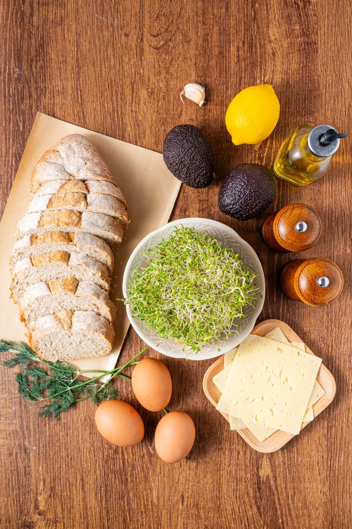 Sliced bread, eggs, cheese, avocado, lemon, sprouts, oil, and seasonings on a wooden table.