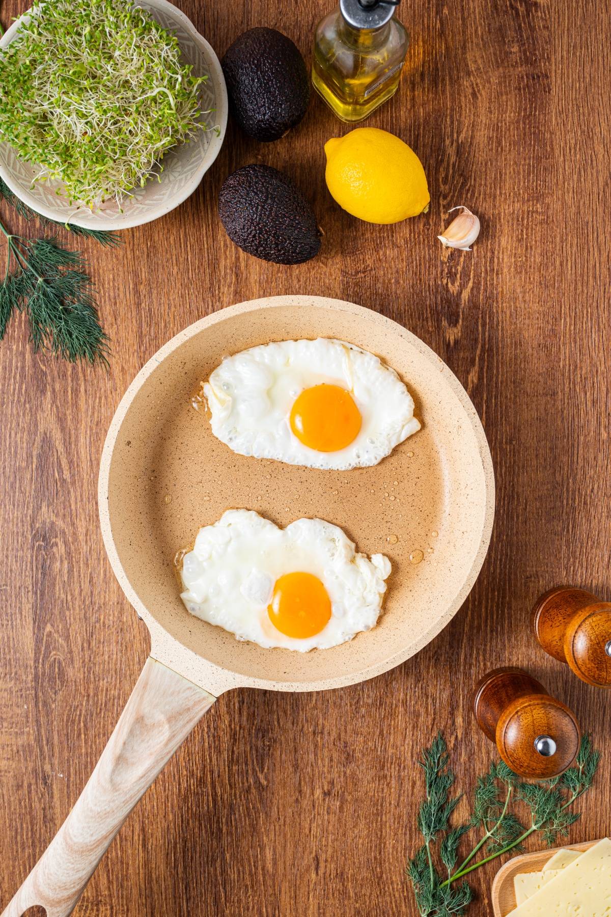Two sunny-side-up eggs cooking in a frying pan on a wooden table with various fresh ingredients around.