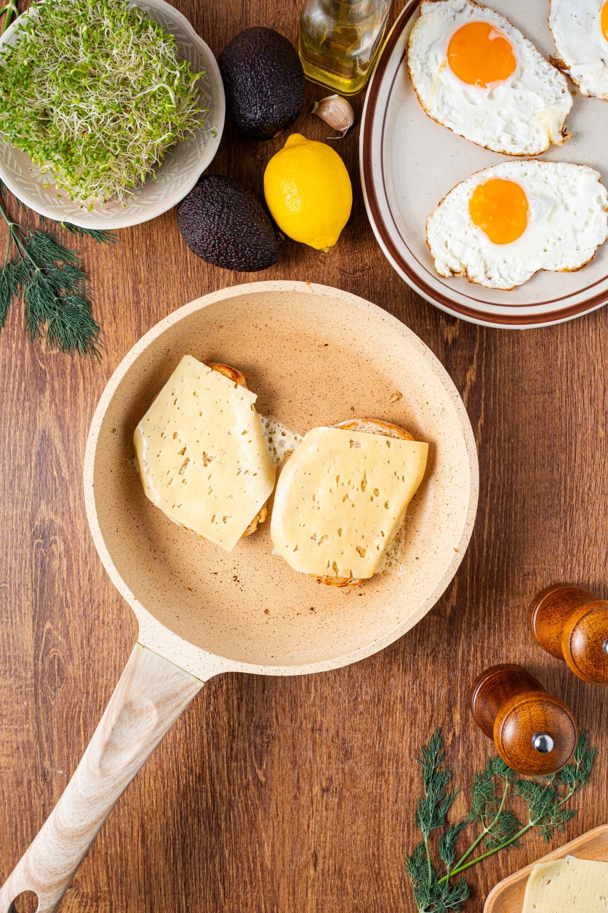 A frying pan with two slices of bread and cheese, surrounded by eggs, avocado, lemon, and herbs.