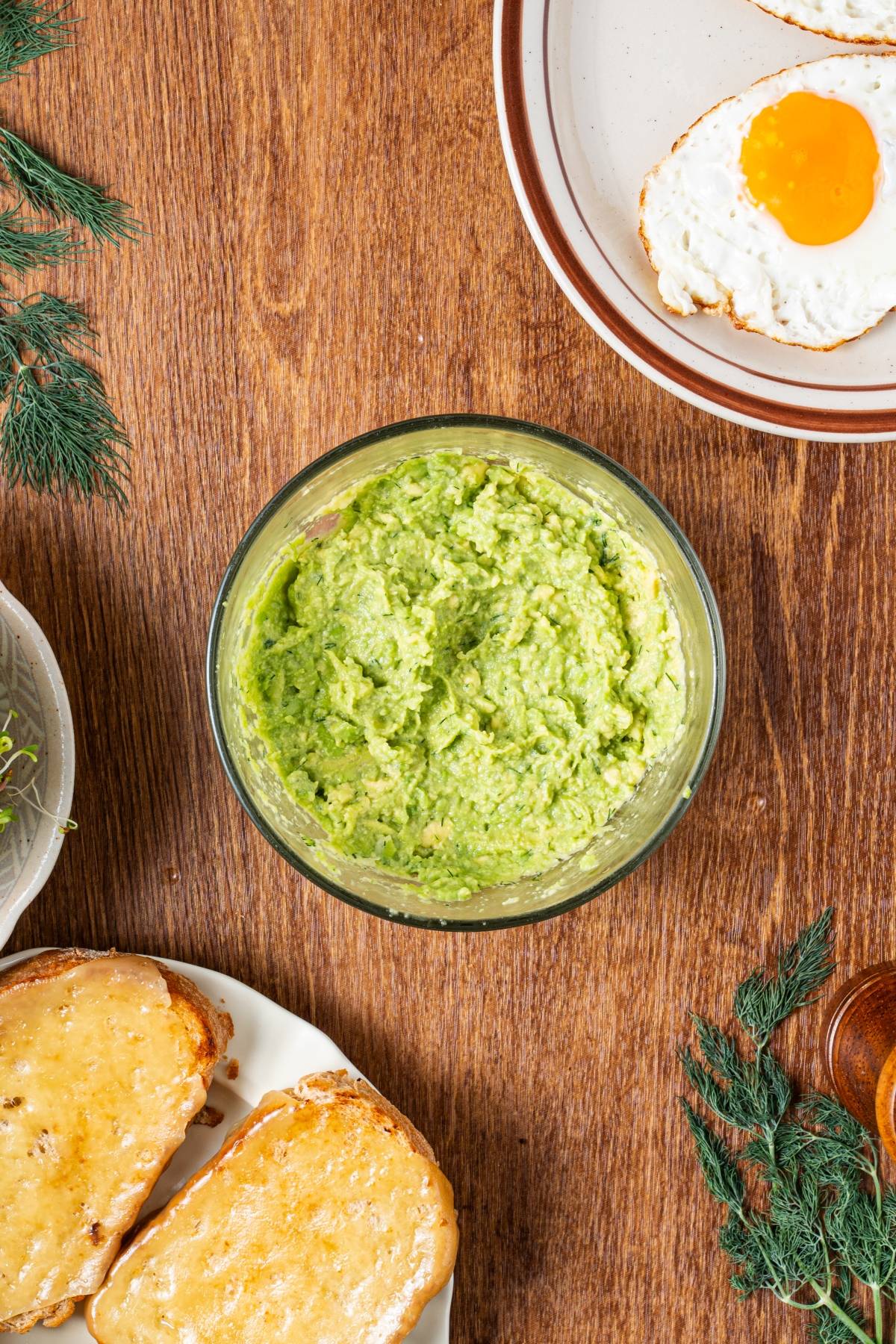 Bowl of mashed avocado, fried egg, and buttered toast on a wooden table, with herbs nearby.
