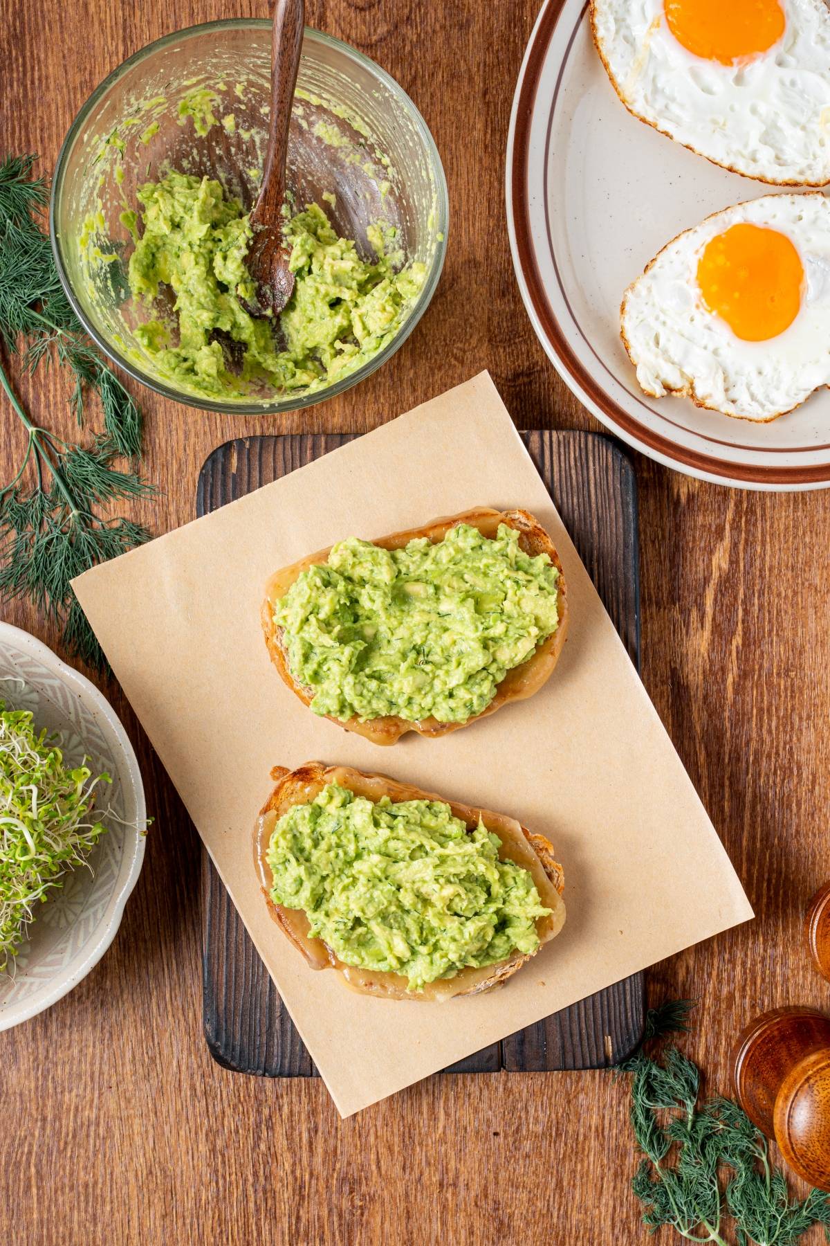 Two slices of bread with mashed avocado, next to fried eggs and a bowl of greens on a wooden table.