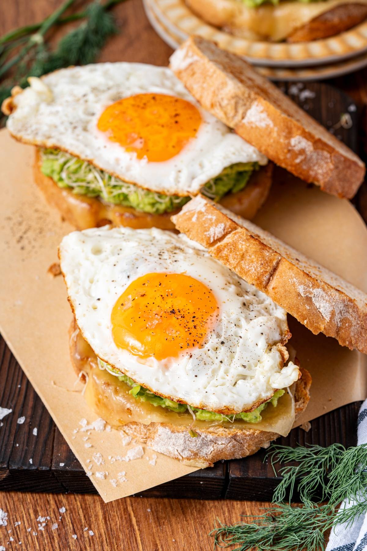 Two sandwiches with fried eggs, avocado, and lettuce on rustic bread, garnished with black pepper.