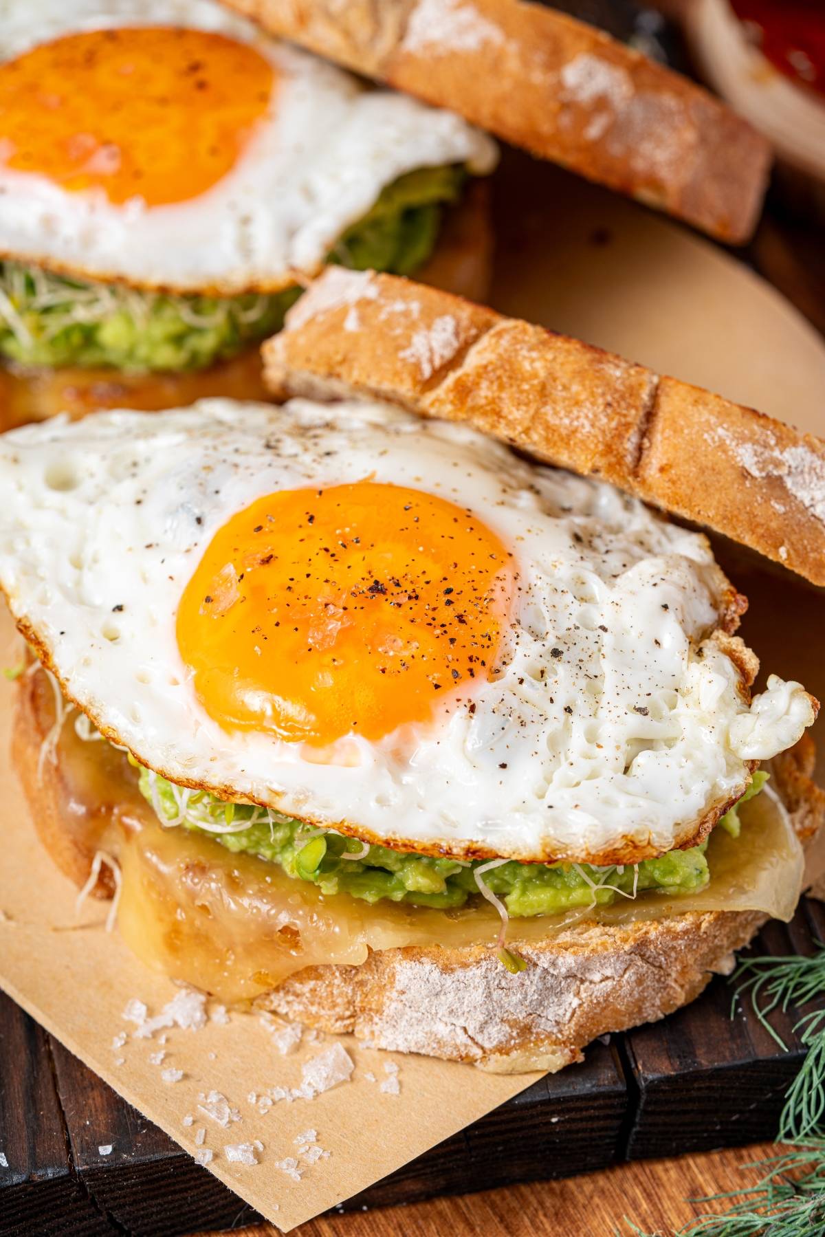 A sandwich with melted cheese, avocado, sprouts, and a sunny-side-up egg on rustic bread.