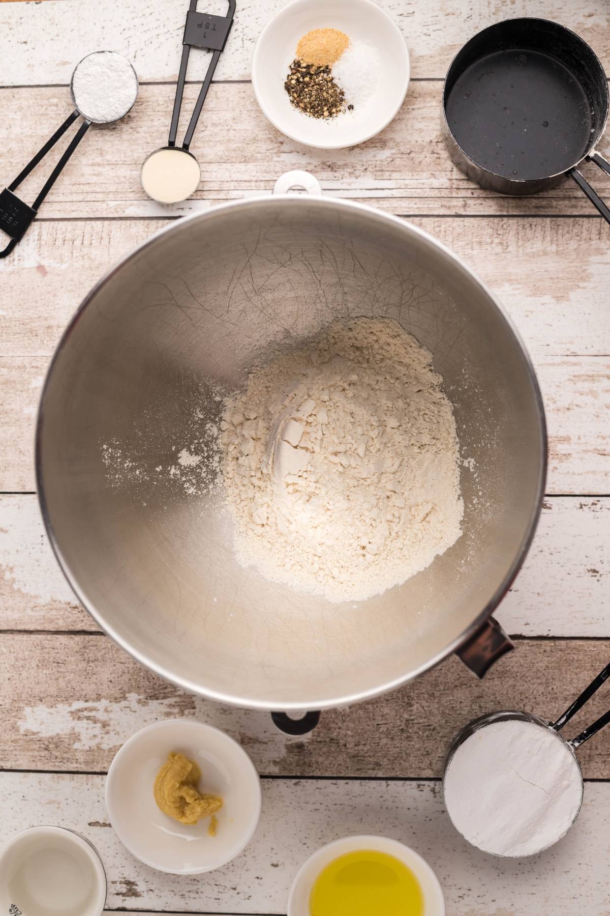 A mixing bowl with flour, perfect for making easy naan recipes like fluffy naan bread without yeast, is surrounded by measuring cups, oil, mustard, spices, and sugar on a wooden table.