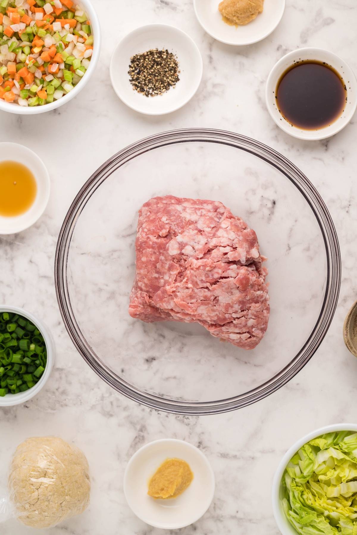A bowl of ground meat surrounded by small bowls of chopped veggies, seasonings, and sauces on a marble surface—perfect ingredients for making gluten free potstickers at home.