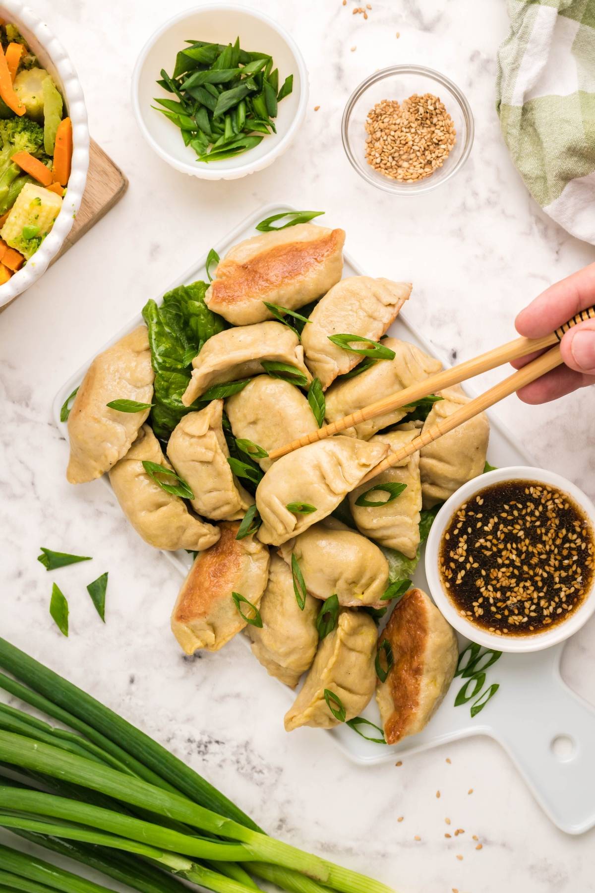 A hand uses chopsticks to pick up a gluten free potsticker from a plate, with sauce, green onions, and vegetables nearby.