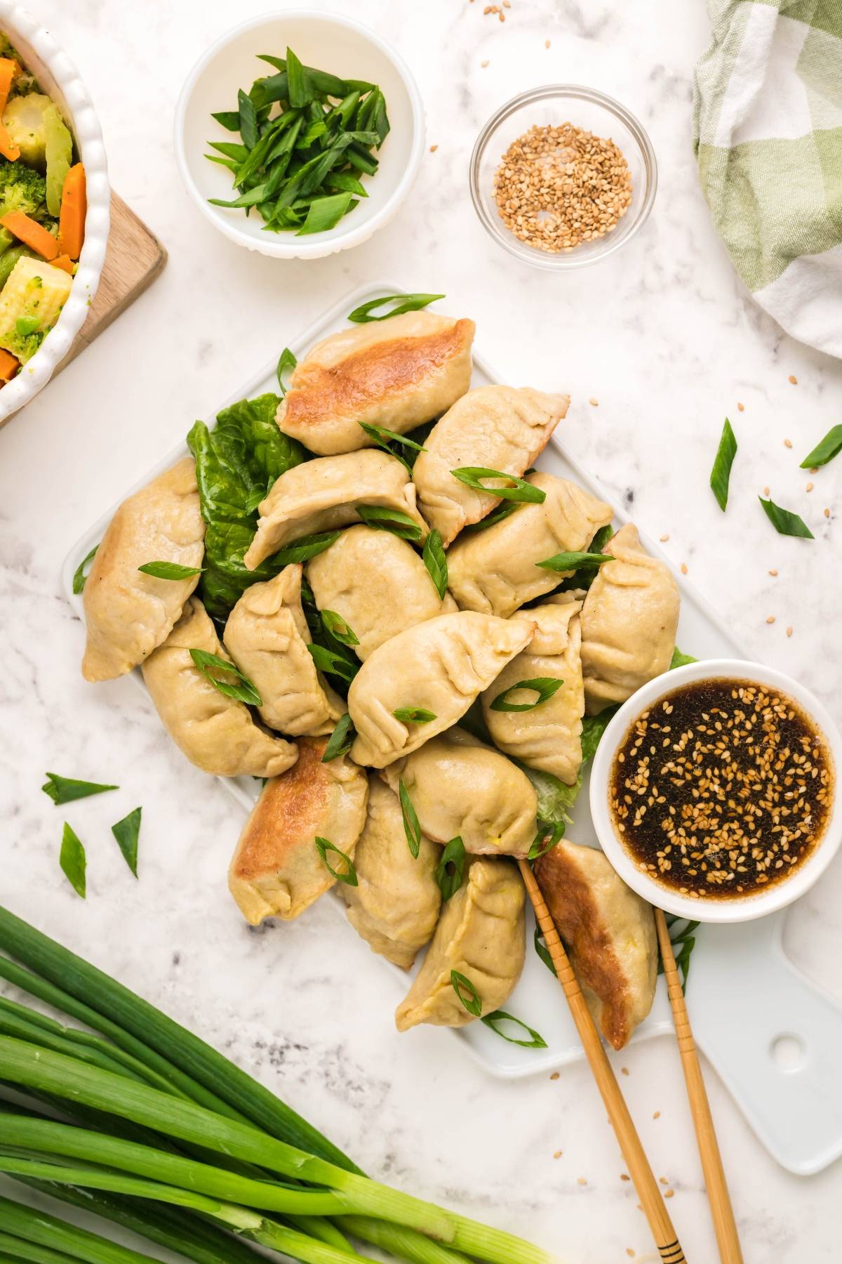A plate of gluten free potstickers with green onions, dipping sauce, chopped scallions, and sesame seeds on a marble surface.