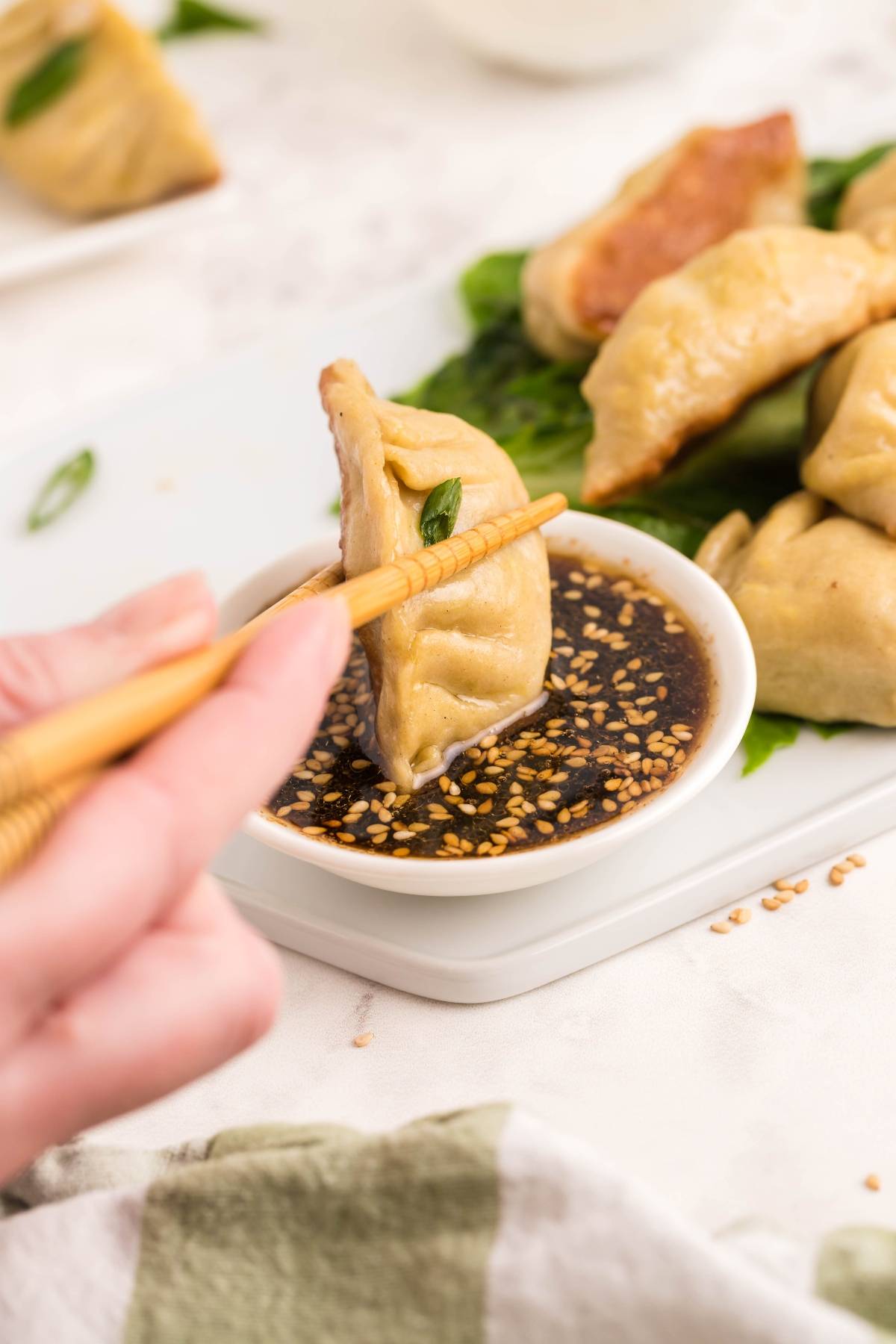 Hand holding chopsticks dips a gluten free potsticker into a small bowl of soy sauce with sesame seeds, with more dumplings nearby.