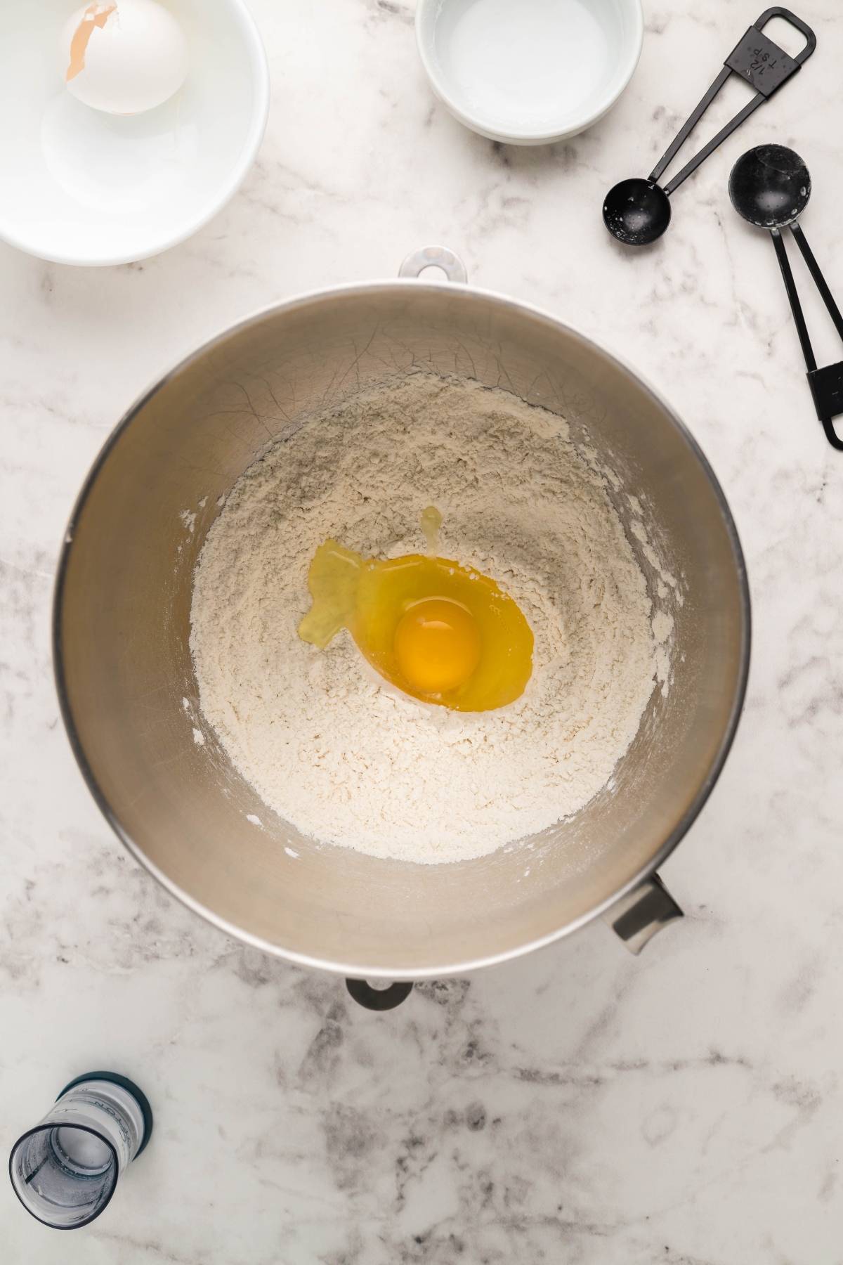 A mixing bowl with flour and a cracked egg, surrounded by baking tools on a marble countertop—perfect for making gluten free potstickers from scratch.