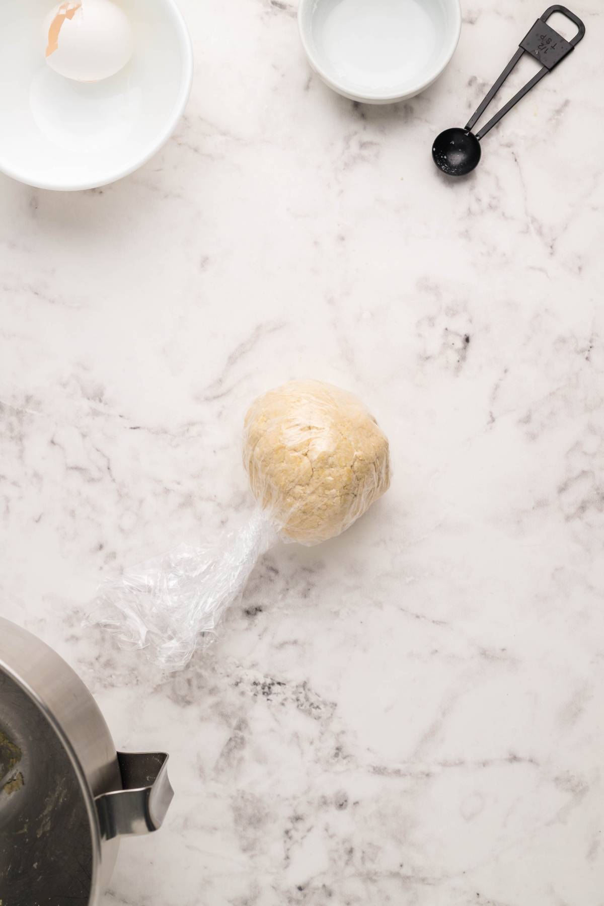 Ball of dough wrapped in plastic wrap on a marble counter, ready for gluten free potstickers, with bowls and measuring spoons nearby.