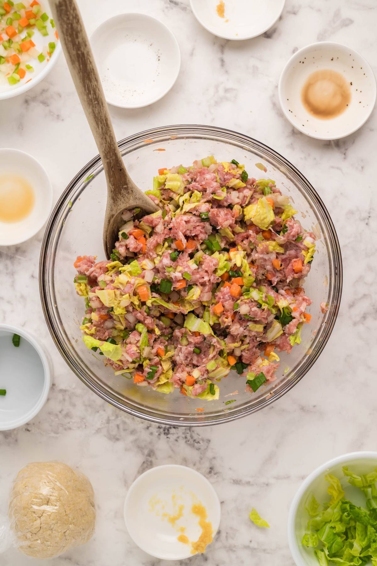 Glass bowl with mixed vegetables and ground meat, surrounded by small dishes and gluten free potstickers on a marble countertop.
