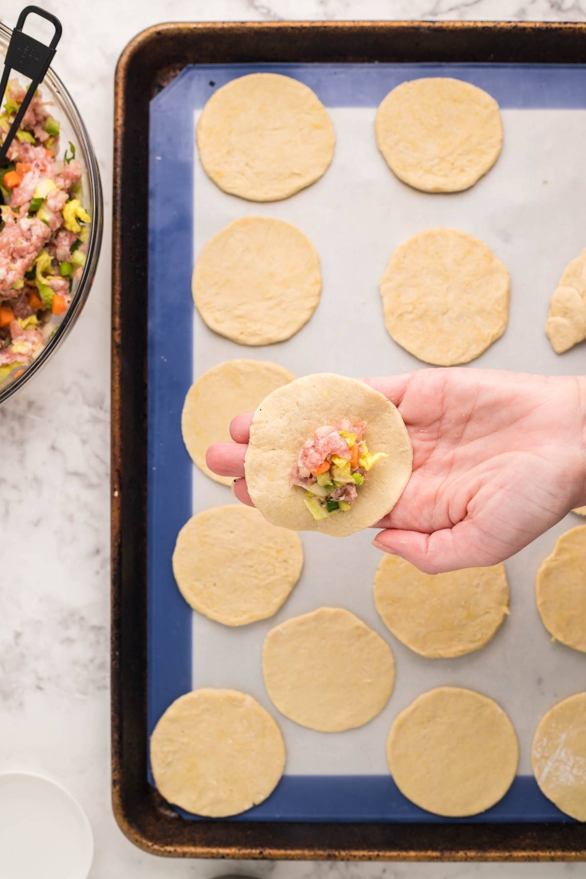 A hand holds a gluten free potsticker dough circle with filling over a baking tray lined with more gluten free potstickers, ready to be cooked.