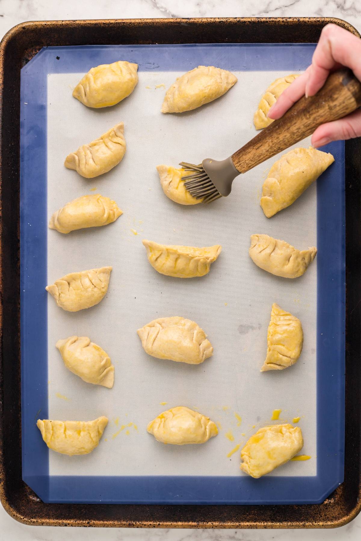 A hand brushes egg wash on unbaked empanadas, arranged on a parchment-lined baking sheet, in a process similar to preparing gluten free potstickers.