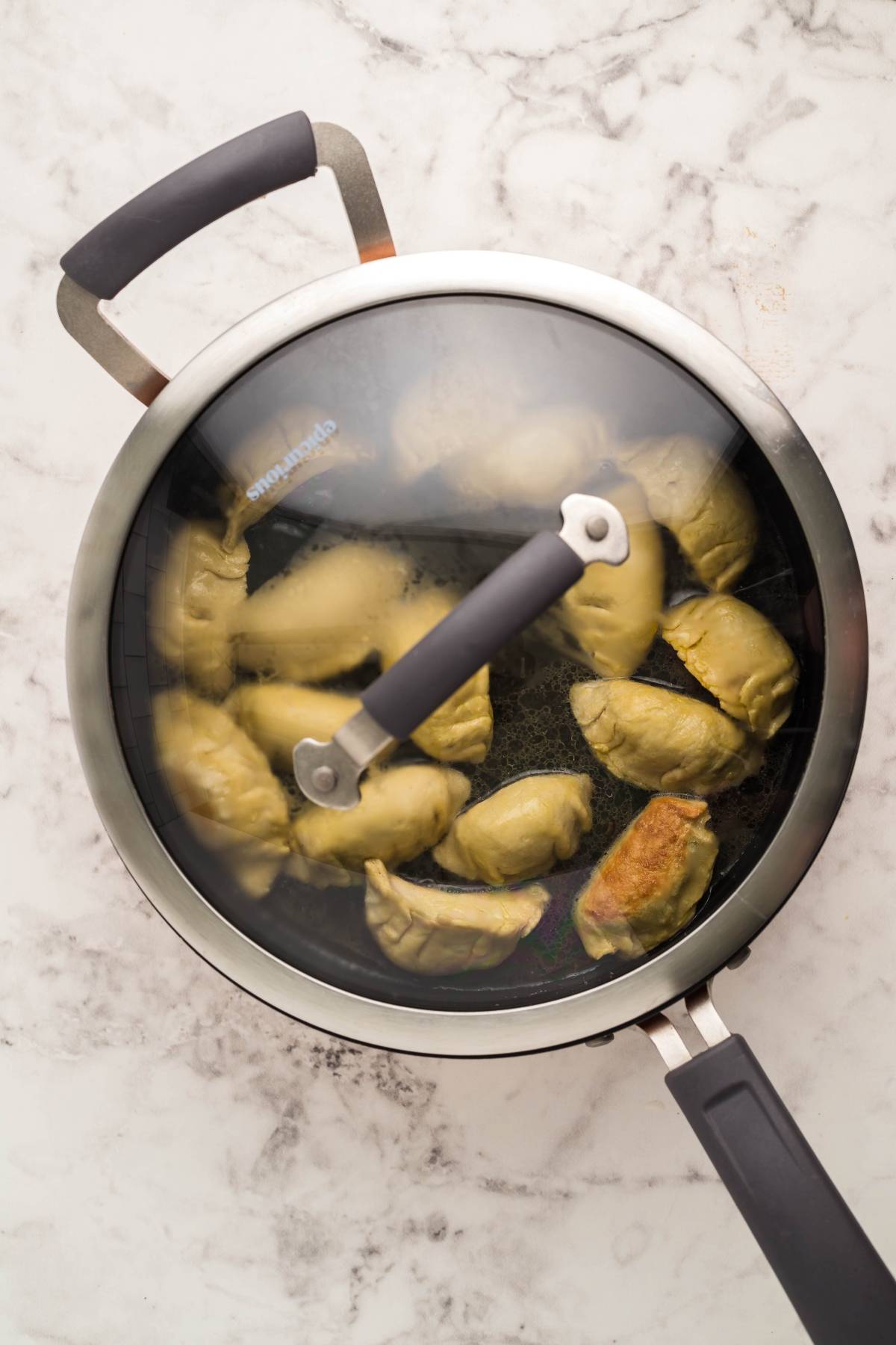 A frying pan with a glass lid cooks gluten free potstickers on a marble countertop.