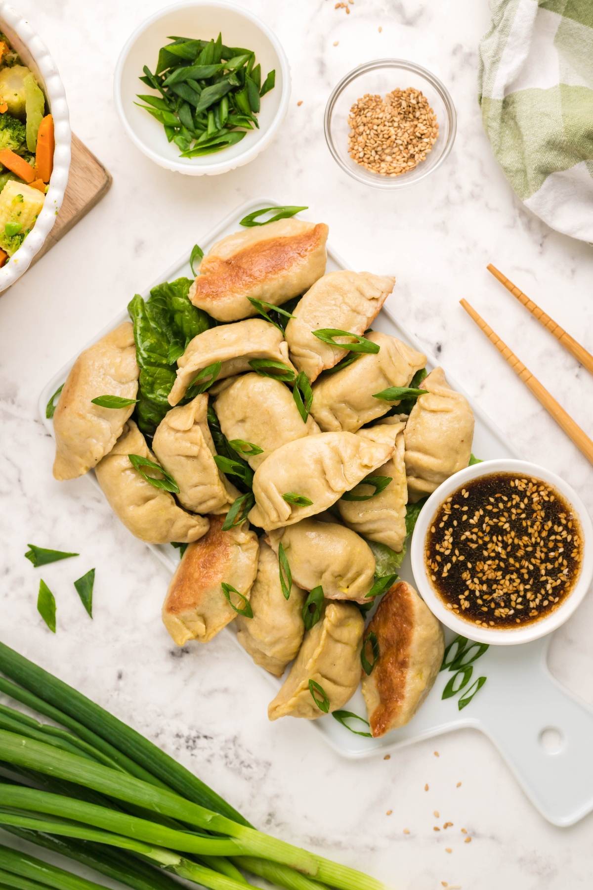 A plate of gluten free potstickers with soy dipping sauce, chopped green onions, and sesame seeds on a marble surface.