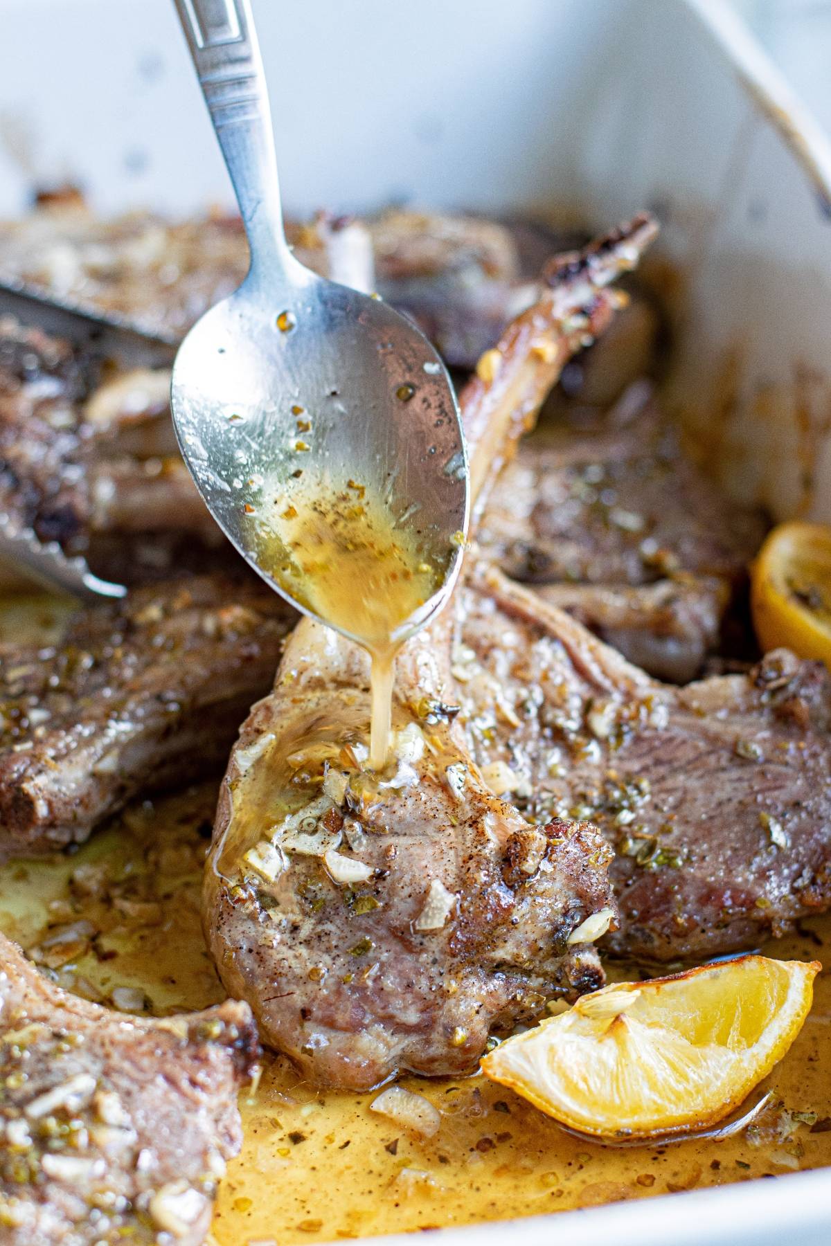 A spoon drizzles marinade over cooked lamb chops in a pan with lemon wedges, evoking the bright flavors of a Greek lemon baked pork chops recipe.