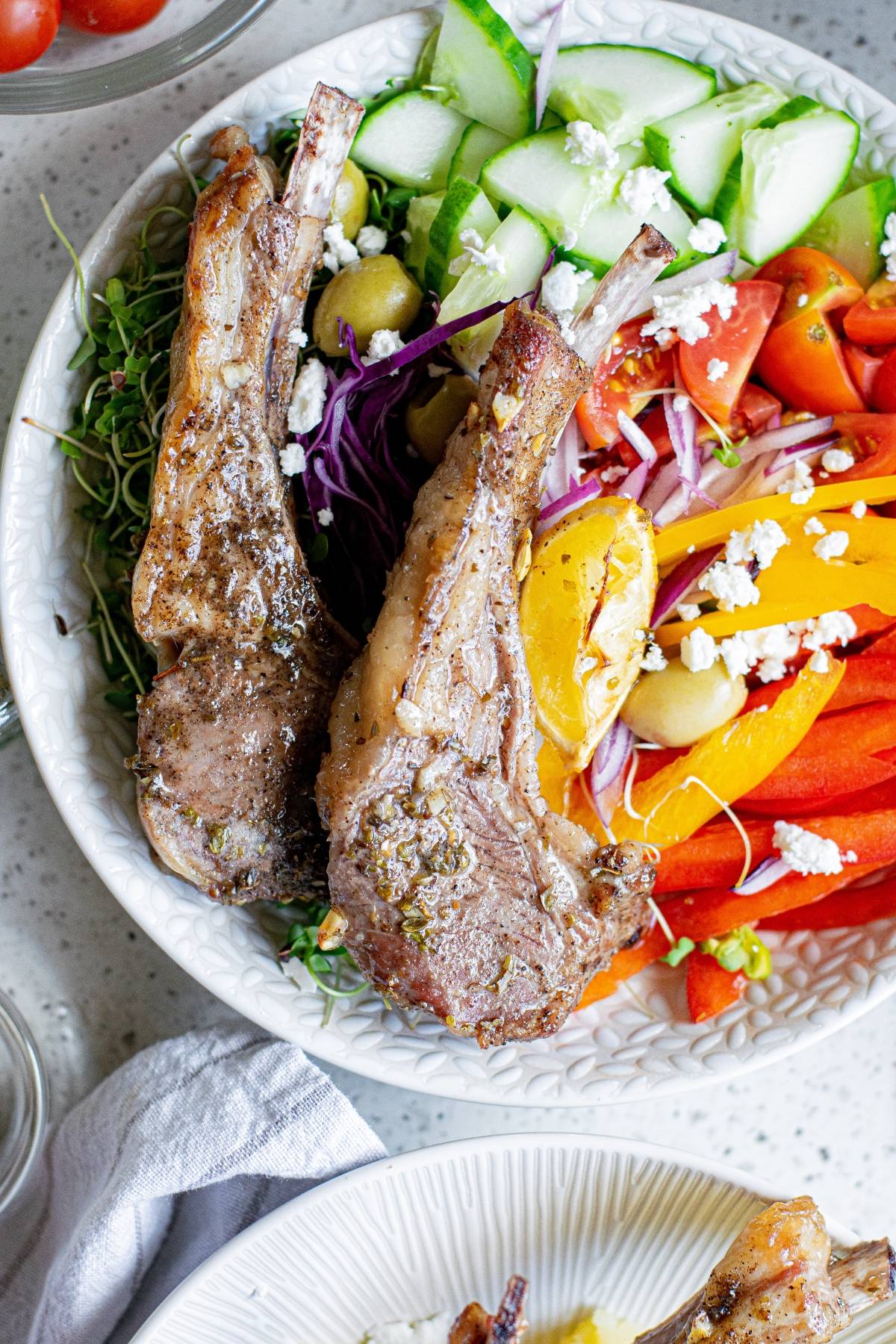A bowl with grilled lamb chops, sliced cucumbers, tomatoes, peppers, olives, and crumbled feta cheese—perfect inspiration for an easy pork recipe or a Greek lemon baked pork chops twist.