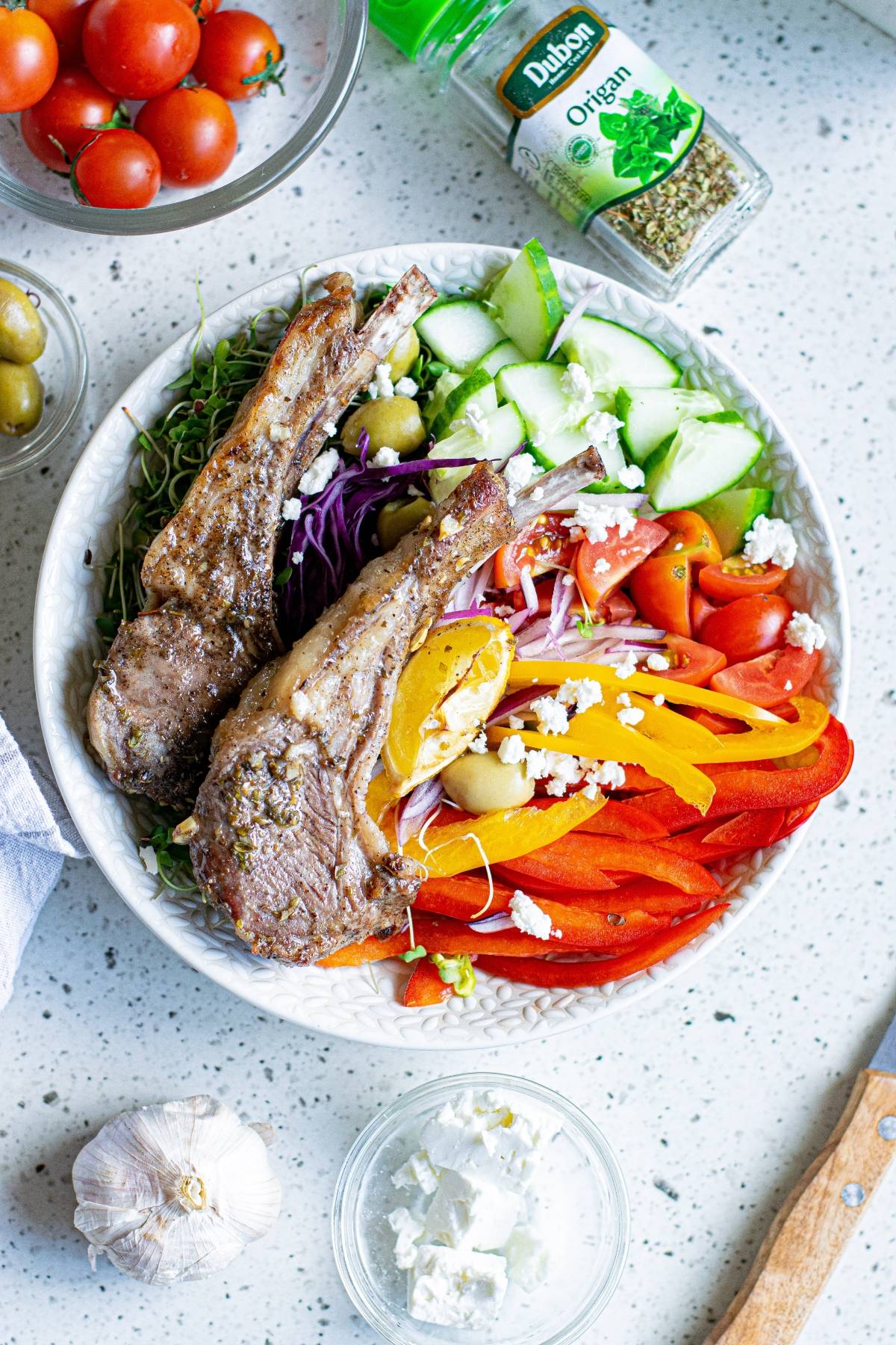 A bowl with Greek pork chops, sliced veggies, olives, sprouts, and feta cheese, on a white counter.