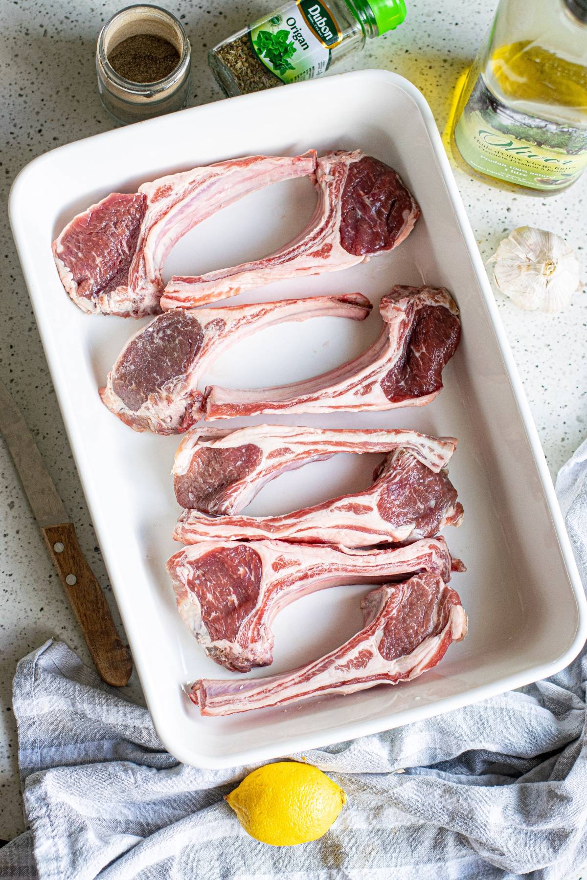 Raw lamb chops arranged in a white baking dish with seasonings, garlic, oil, and a lemon nearby—perfect inspiration for an easy pork recipe like Greek lemon baked pork chops.