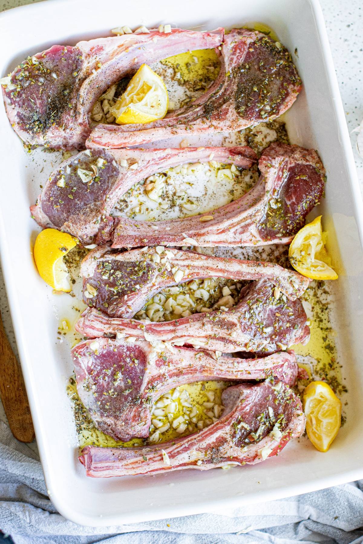 Raw lamb chops in a white baking dish with chopped garlic, herbs, and lemon wedges—similar to an easy Greek pork chops recipe, but with tender lamb for a Mediterranean twist.