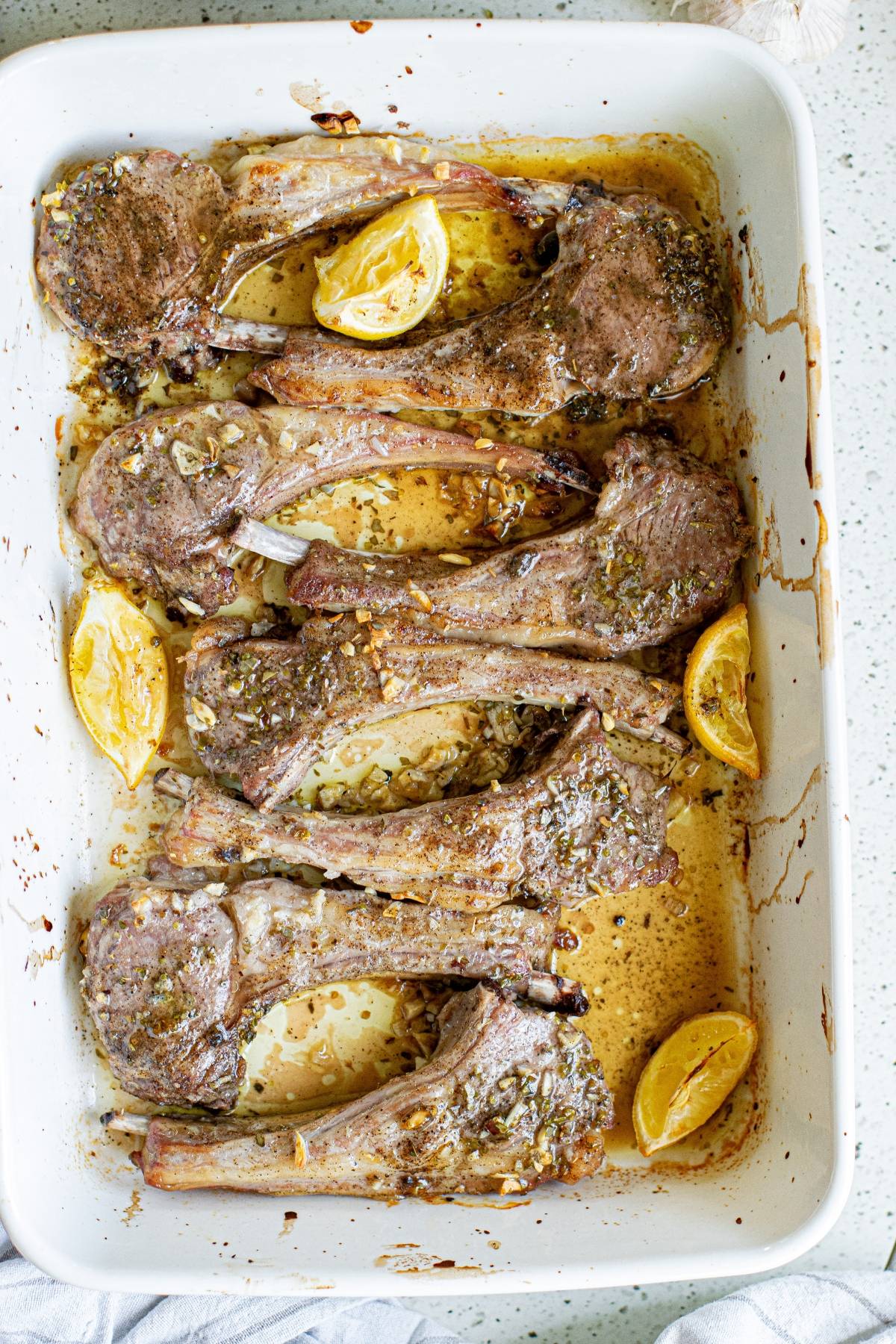 Six cooked lamb chops with herbs and lemon wedges in a white baking dish on a speckled countertop, inspired by Greek lemon baked pork chops.
