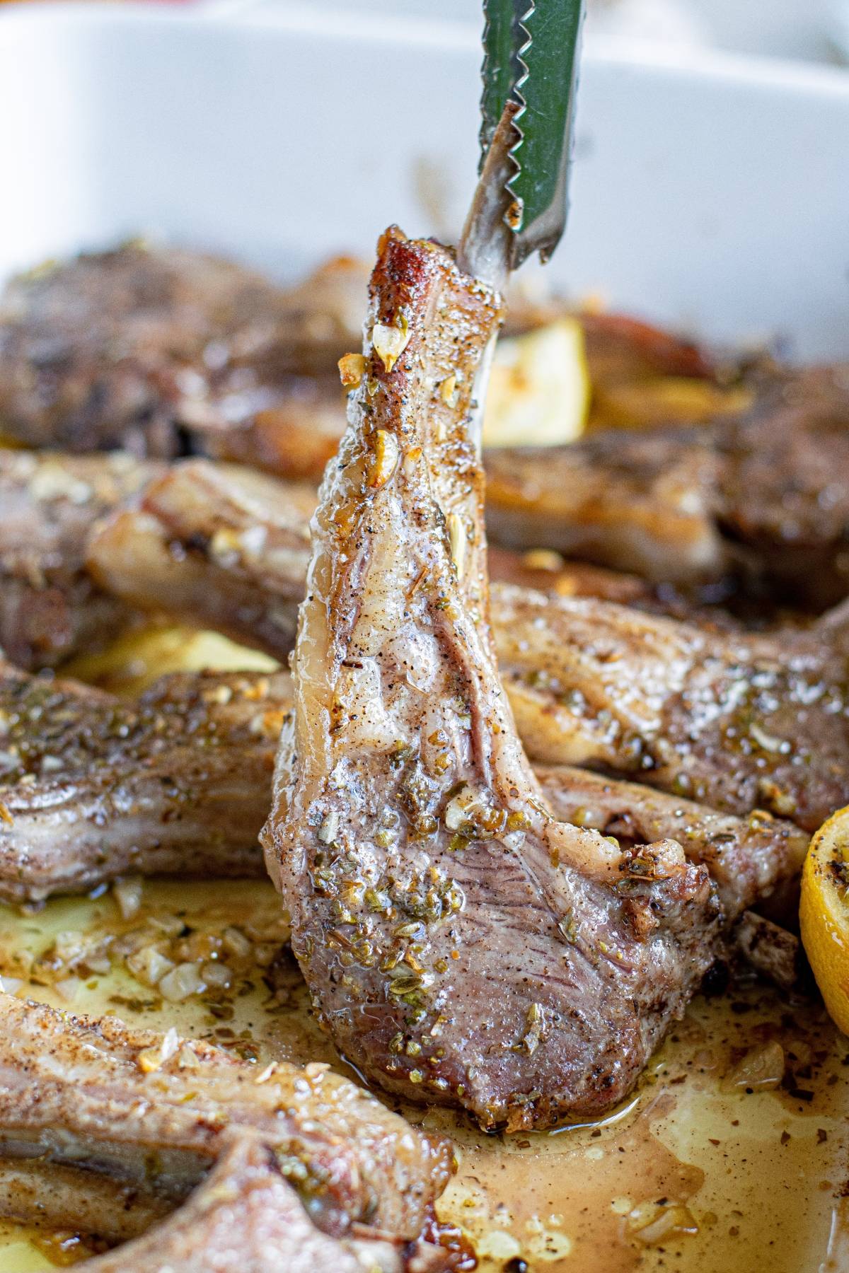 A pair of tongs holding a seasoned, cooked Greek pork chop above a tray of more chops.
