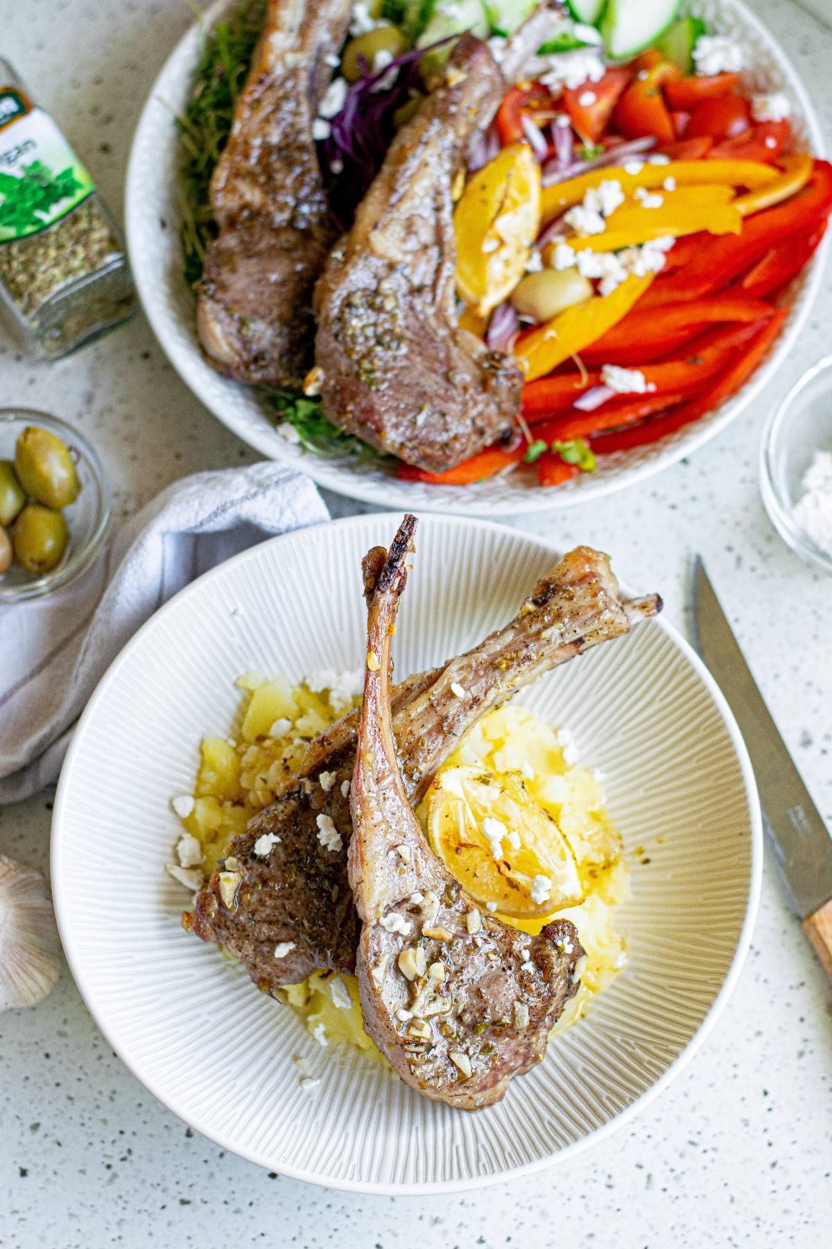 Two grilled lamb chops on mashed potatoes with a lemon wedge, served beside a colorful Greek salad—an elegant alternative to the classic Greek pork chops or any baked pork chops recipe.
