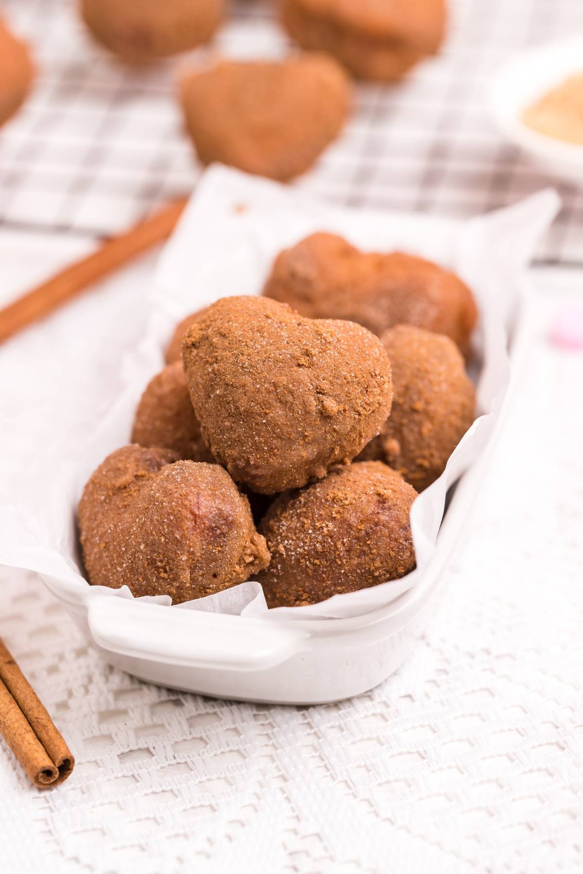 Heart-shaped brown cookies in a white dish, with cinnamon sticks and more cookies in the background—perfect inspo for your next mini doughnut recipe or a heart shaped doughnut hole twist using a clever cinnamon roll hack.