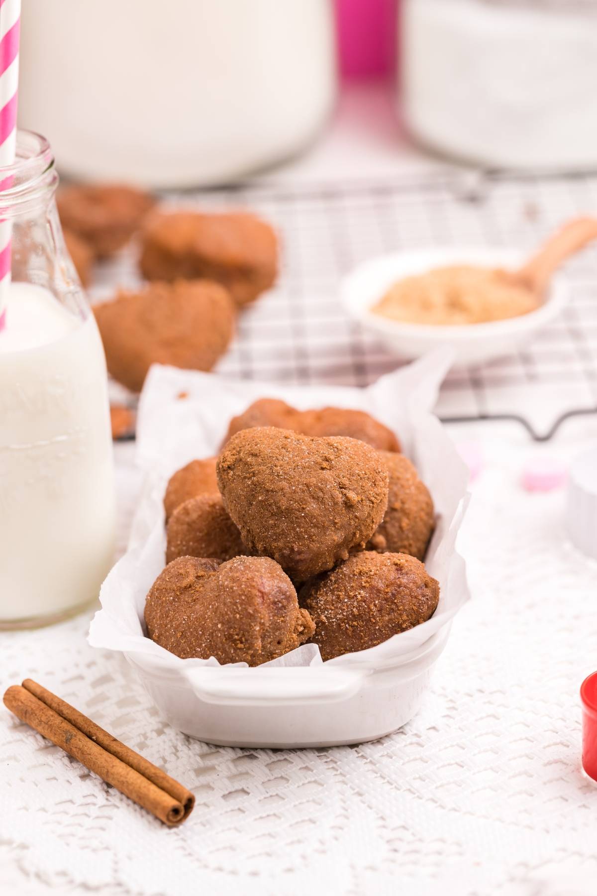 Heart-shaped brown cookies in a white dish, with a bottle of milk and cinnamon sticks nearby—perfect inspiration for your next mini doughnut recipe or cinnamon roll hack.