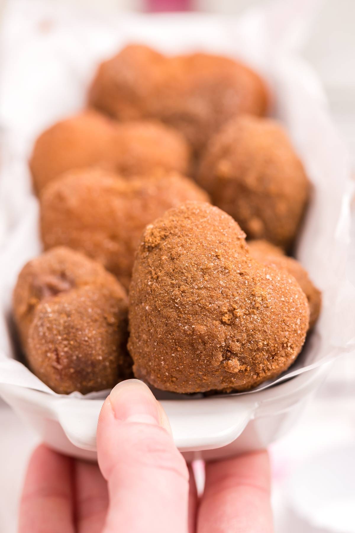 A hand holds a dish of heart-shaped doughnut holes, cinnamon-coated and lined up closely together—perfect for trying out a fun mini doughnut recipe.
