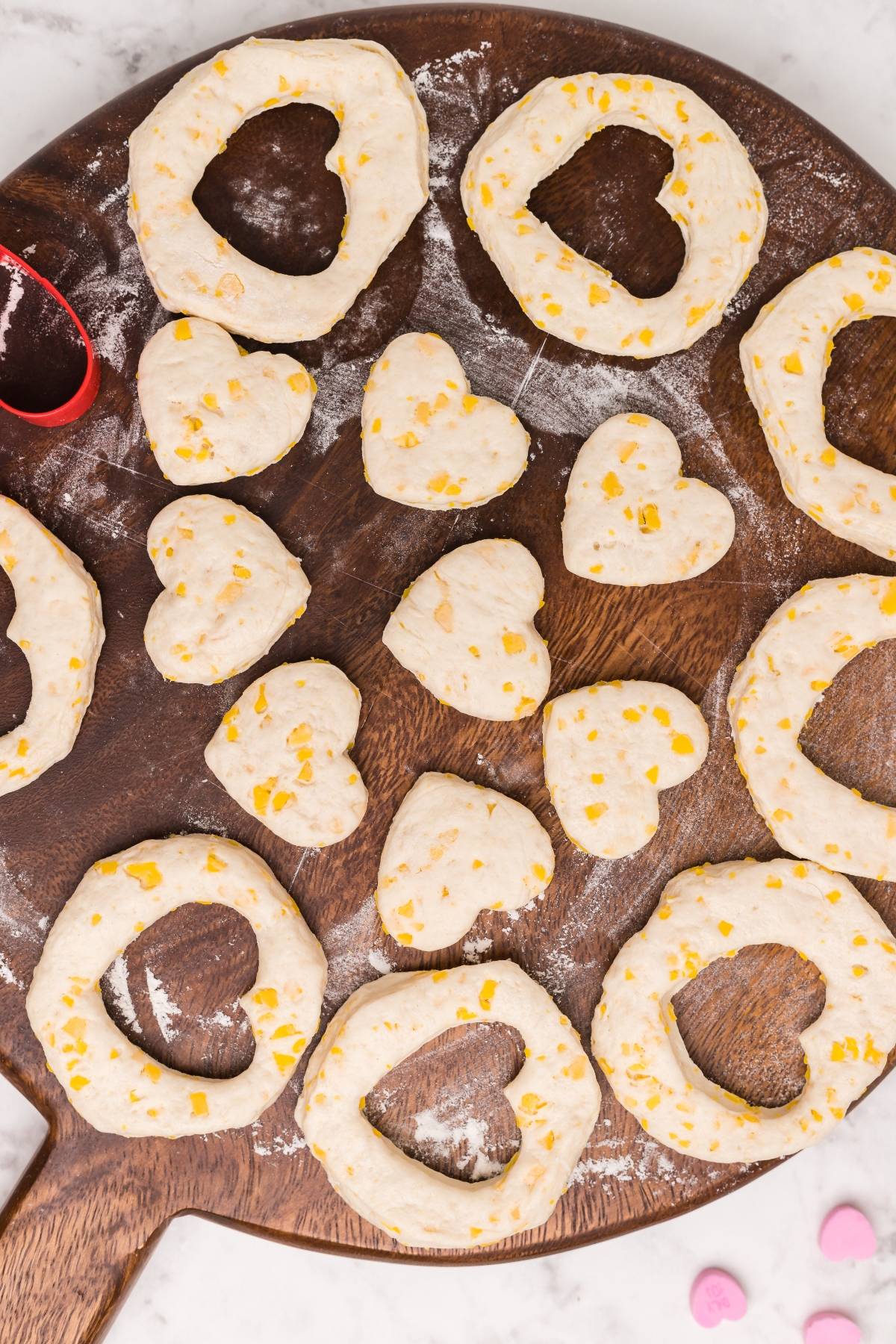 Unbaked heart-shaped and ring-shaped dough pieces with cheese on a floured wooden board—perfect for trying a mini doughnut recipe or creating adorable heart shaped doughnut holes.