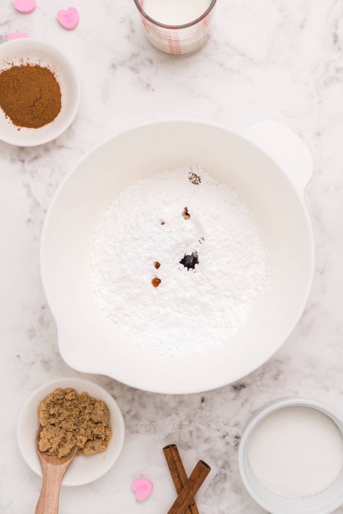 Mixing bowl with powdered sugar, vanilla, and spices, surrounded by brown sugar, cinnamon, and milk on a marble surface—the perfect setup for a mini doughnut recipe or trying a fun cinnamon roll hack.