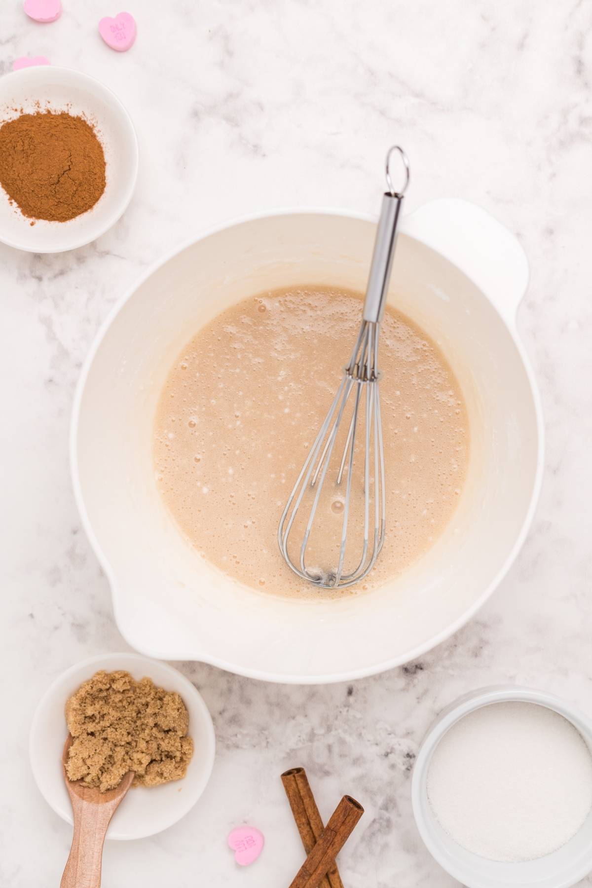 A mixing bowl with batter and a whisk, surrounded by cinnamon, brown sugar, white sugar, and cinnamon sticks—perfect for trying a mini doughnut recipe or testing your favorite cinnamon roll hack.