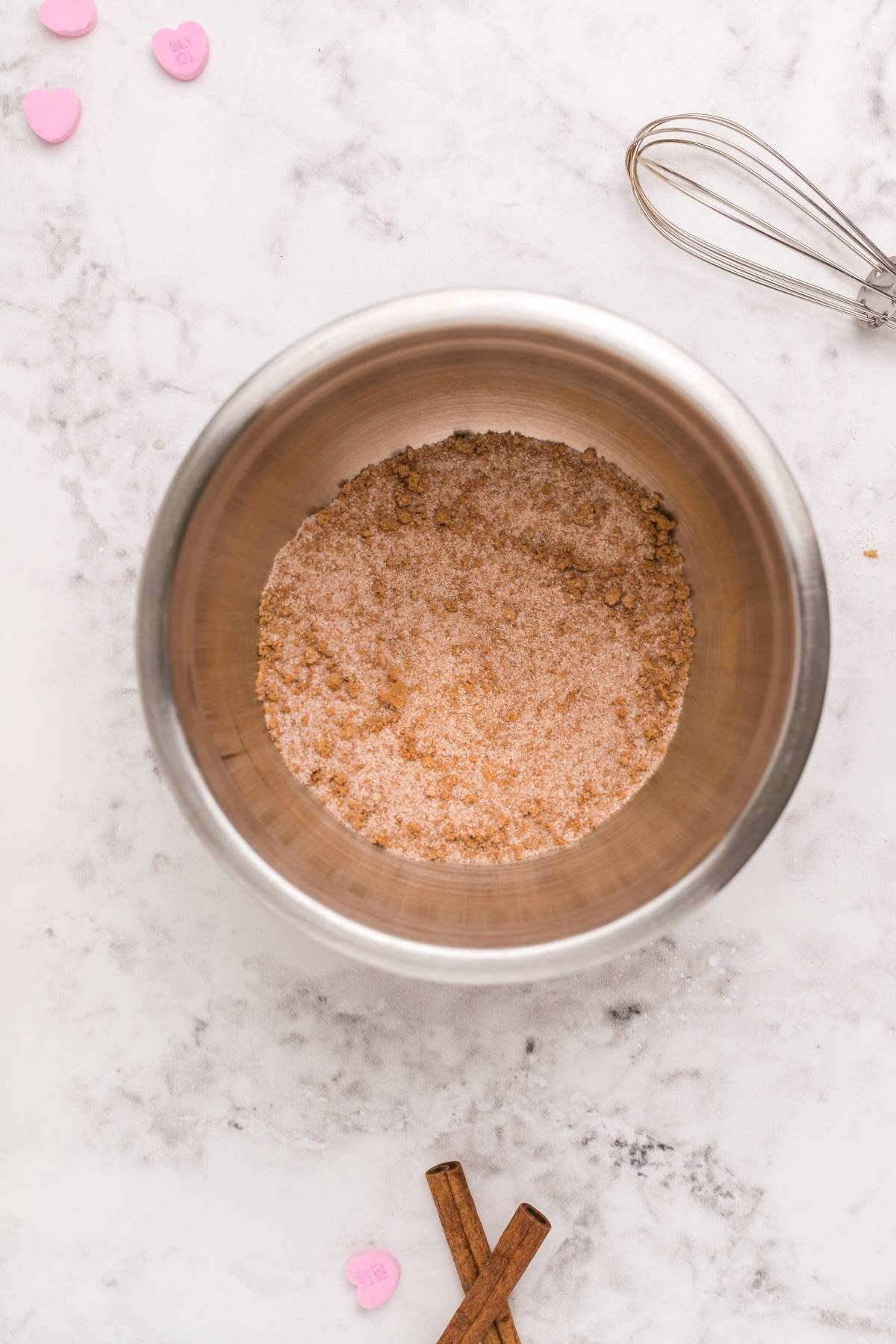 A metal bowl with cinnamon sugar on a marble surface, perfect for your mini doughnut recipe, with a whisk, candy hearts, and cinnamon sticks nearby.