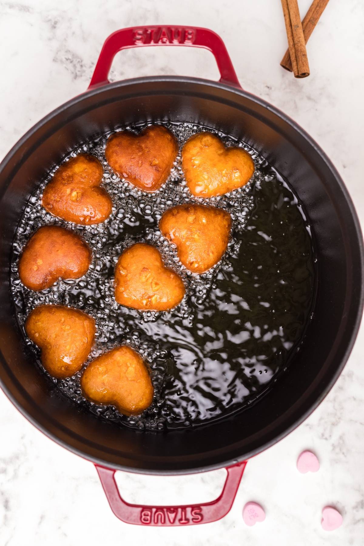 Heart-shaped doughnuts frying in oil inside a red Dutch oven, with cinnamon sticks on the side—perfect inspiration for your next mini doughnut recipe.